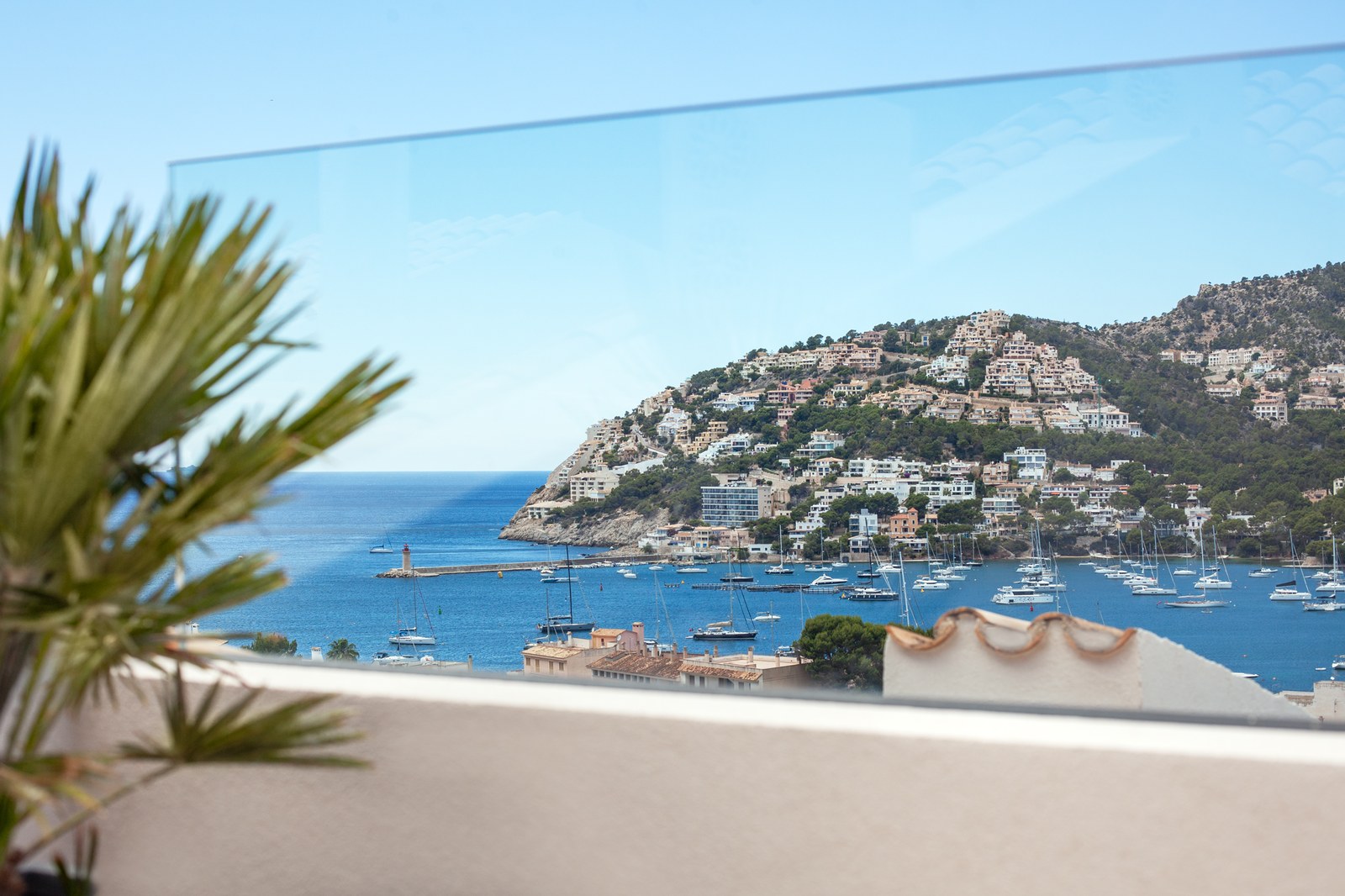 View through a glass balcony railing of a Mediterranean harbor filled with boats and a hillside town.