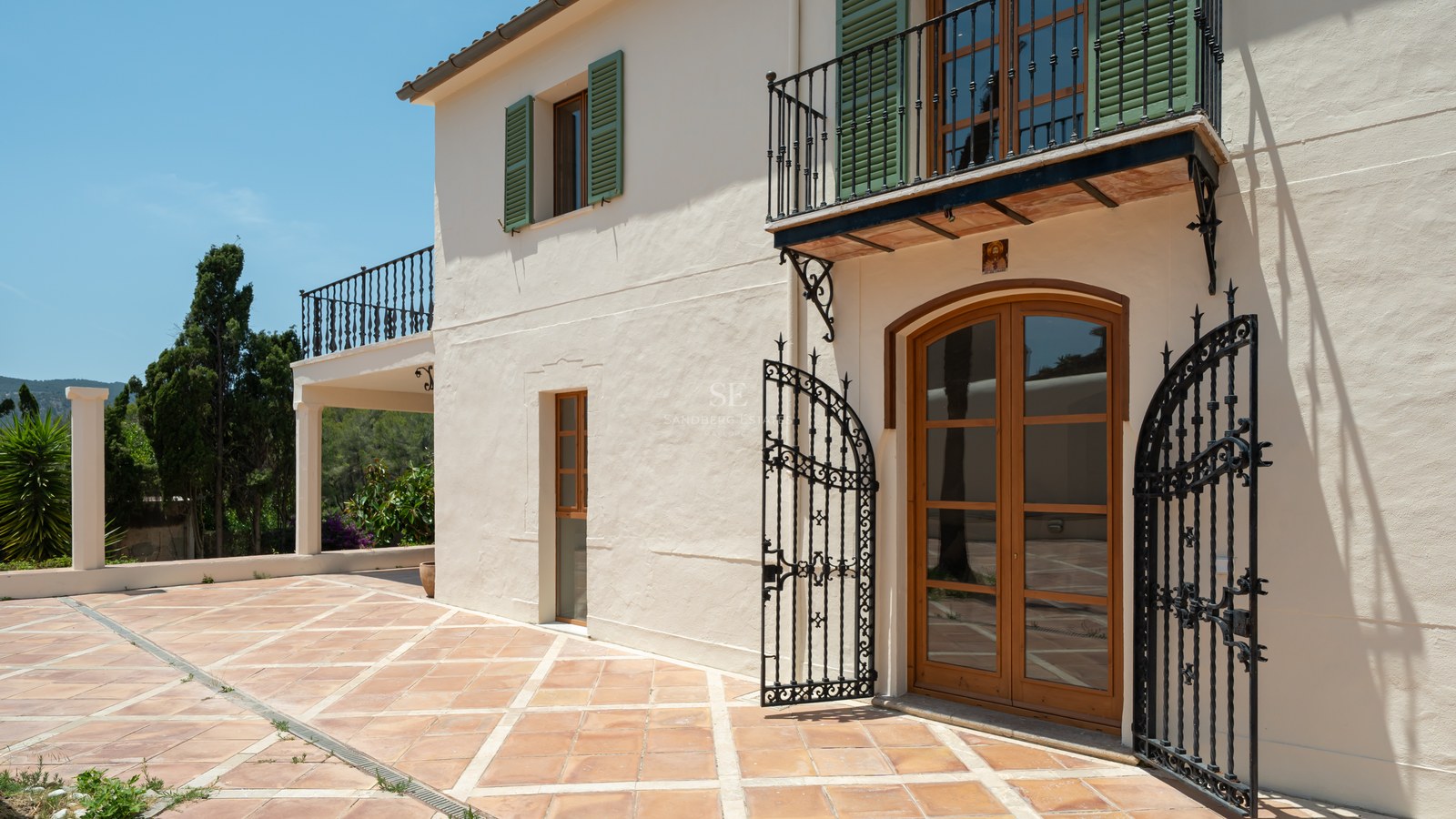 Cream stucco villa with green shutters, wrought iron balcony, and terracotta terrace under a clear blue sky.