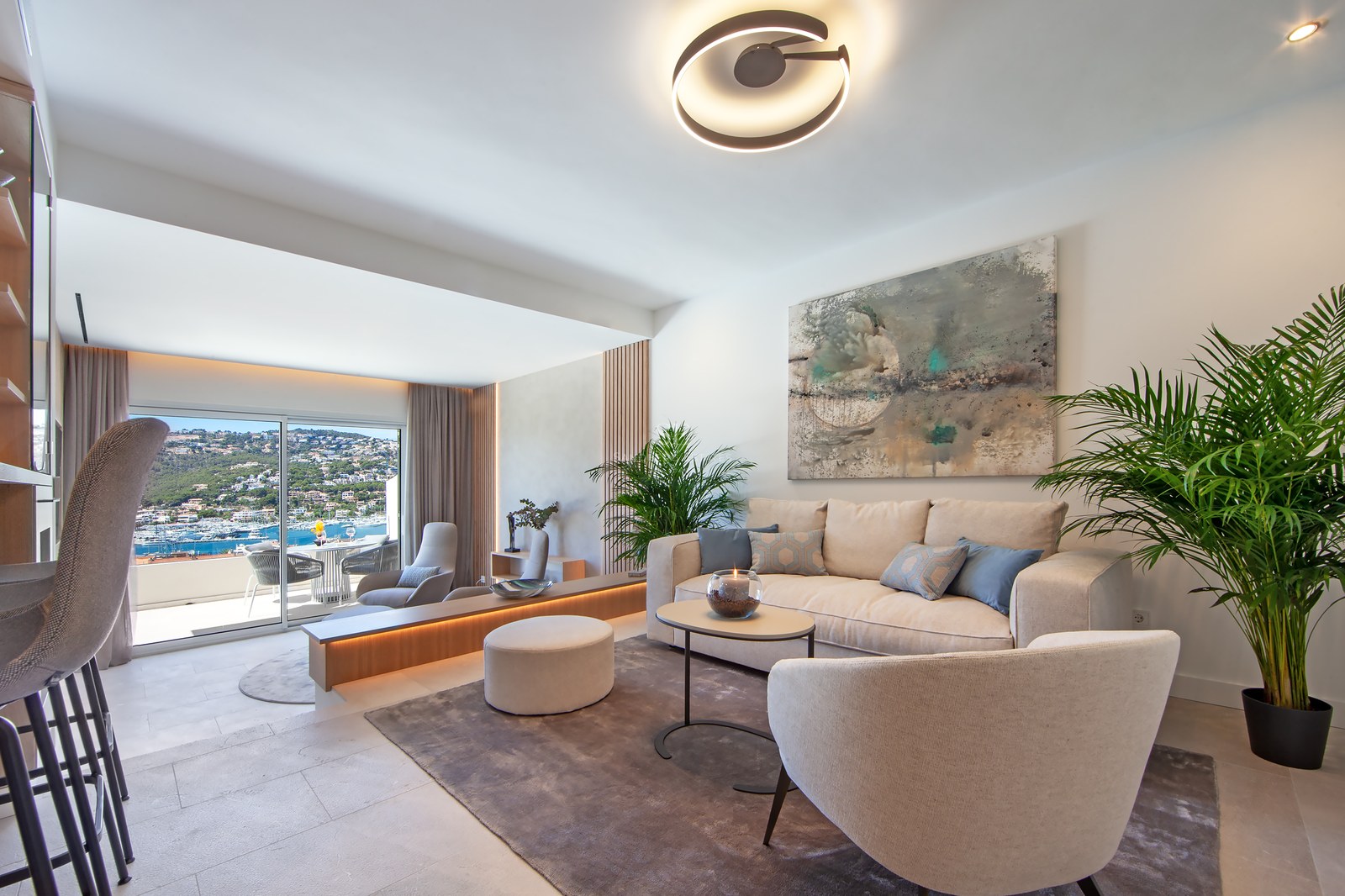 Bright modern living room with cream furniture, large windows overlooking the sea, and lush indoor plants.