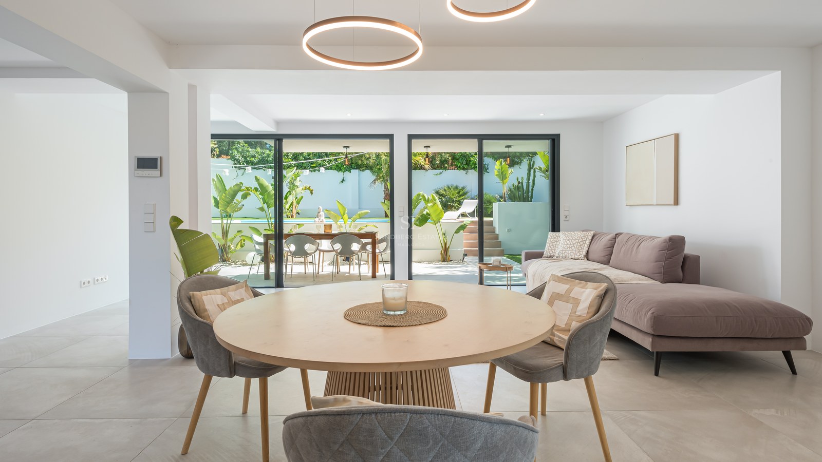 Contemporary living and dining room with circular LED lighting, grey sofa, and views to a pool and tropical garden.