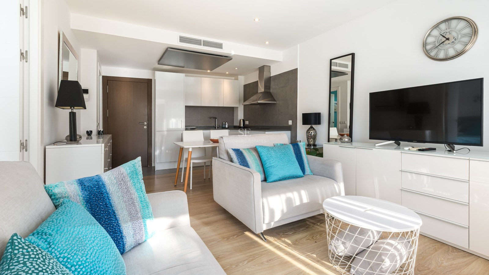 Bright living room with grey armchair, teal cushions, TV console, and modern kitchen in the background.