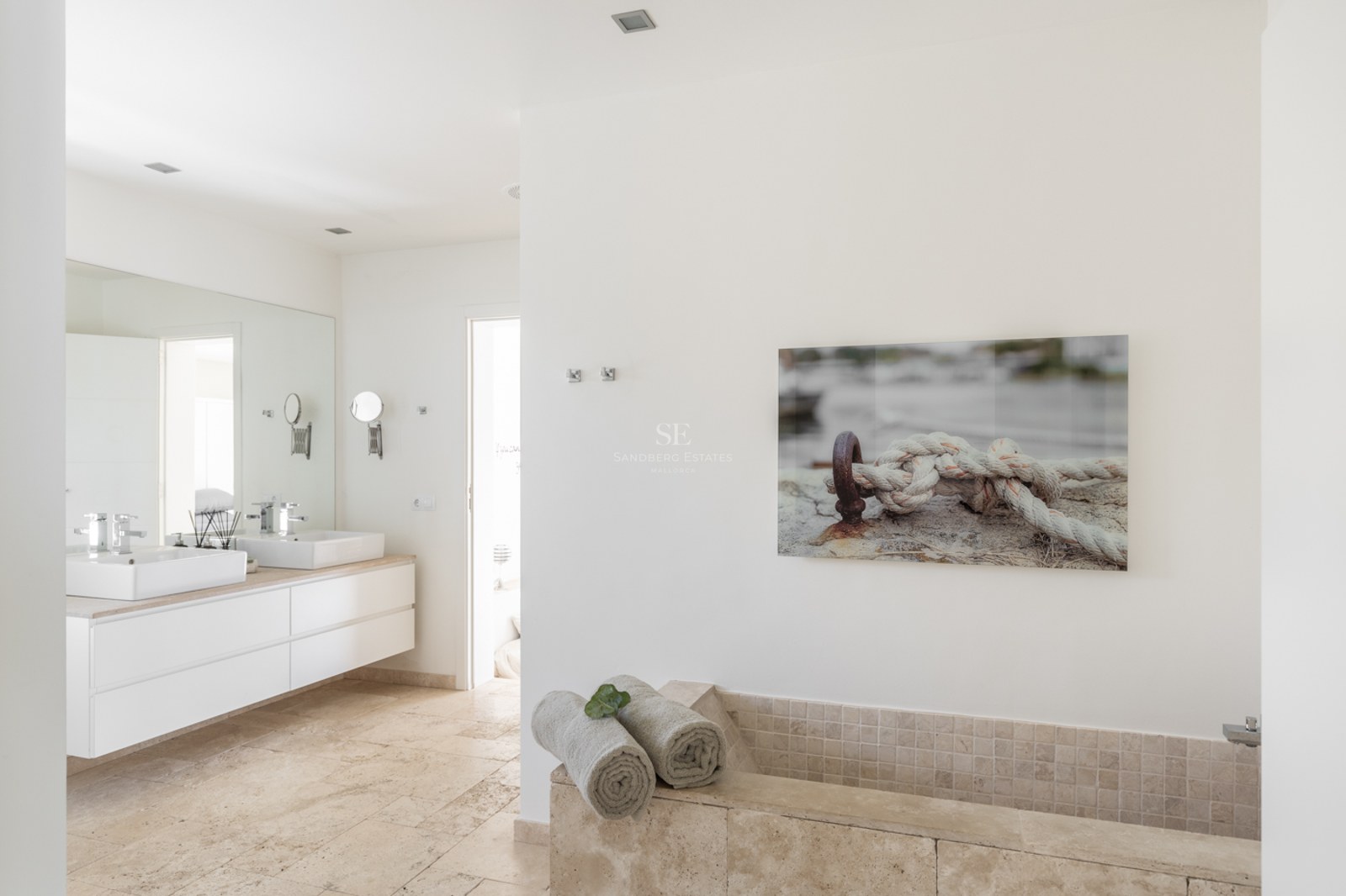 Salle de bains lumineuse avec double vasque suspendue, sol en travertin et baignoire intégrée en pierre.