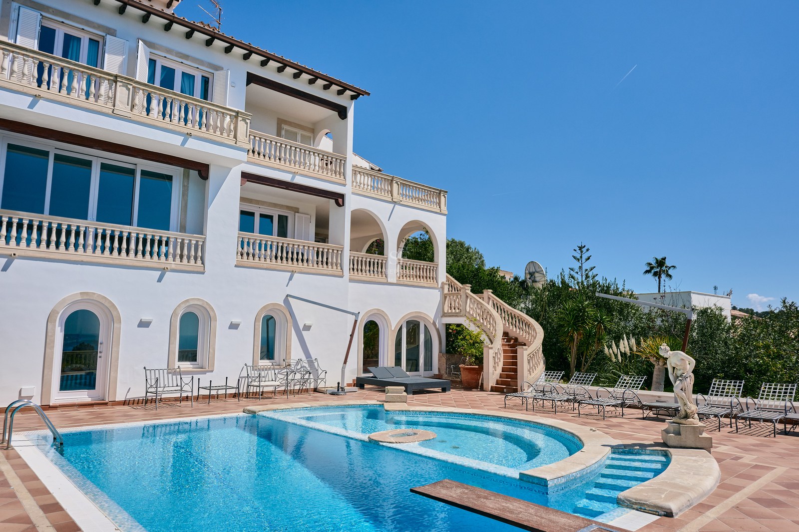Large white multi-level villa with a curved blue swimming pool, stone balustrades, and a terracotta sun deck.