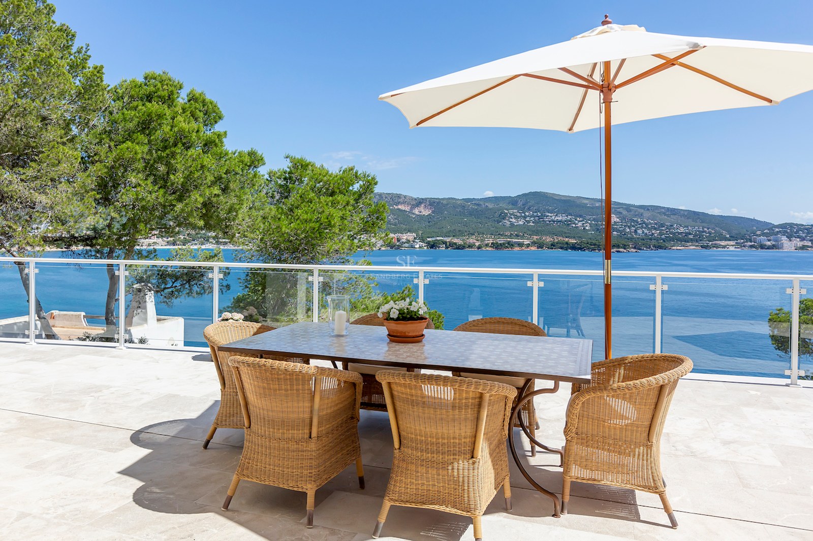 Outdoor dining area with wicker chairs and white umbrella on a stone terrace overlooking the blue sea and mountains.
