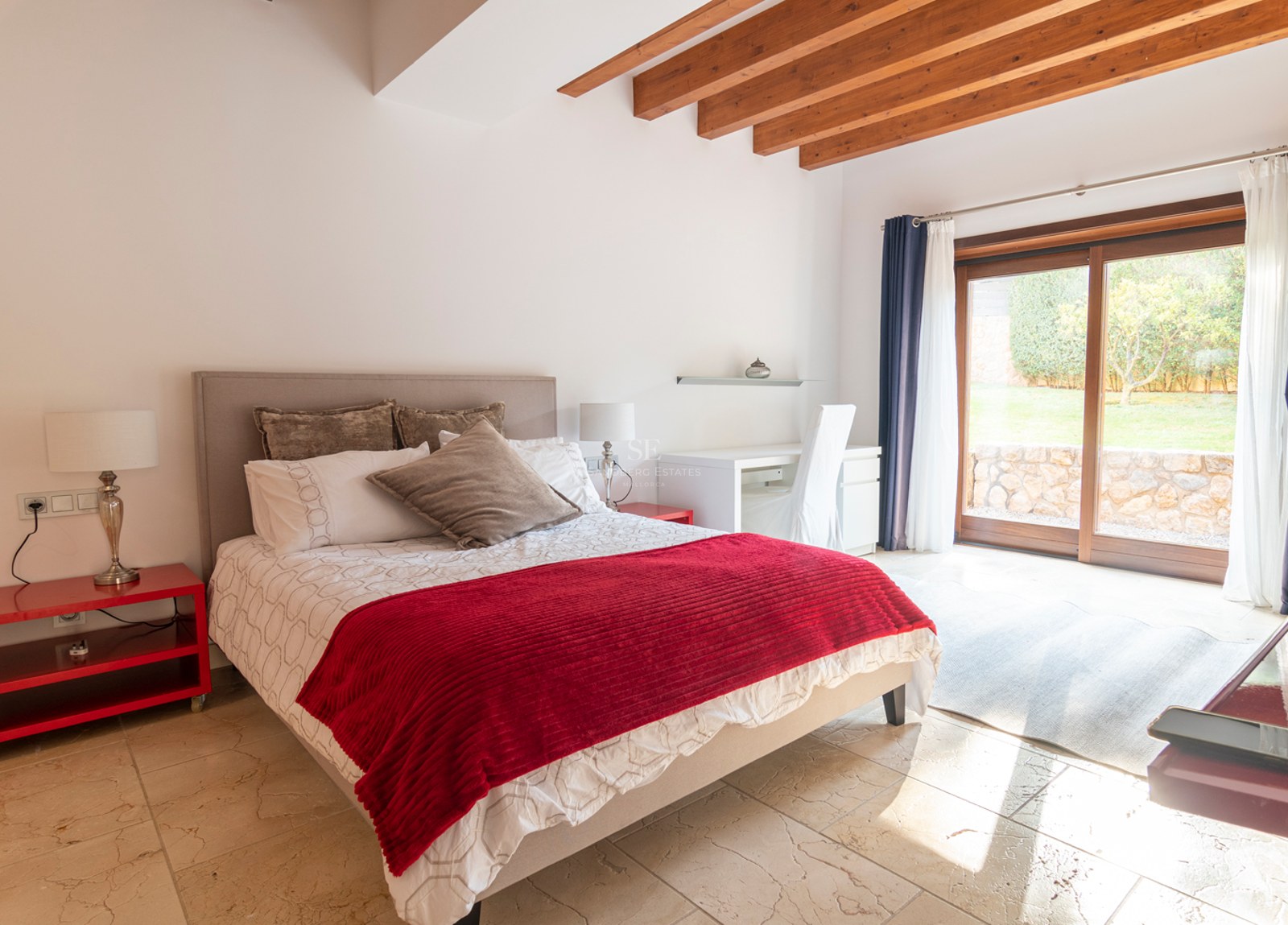 Chambre lumineuse avec accents rouges, sol en pierre, poutres en bois et baies vitrées donnant sur le jardin.