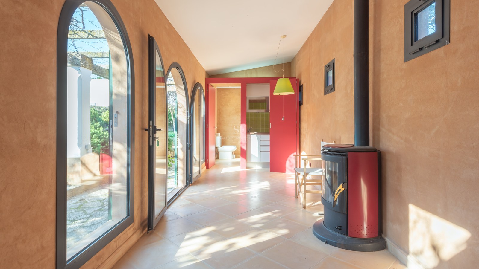 Sunlit room with terracotta walls, arched glass doors, a red wood-burning stove, and a view to an outdoor patio.