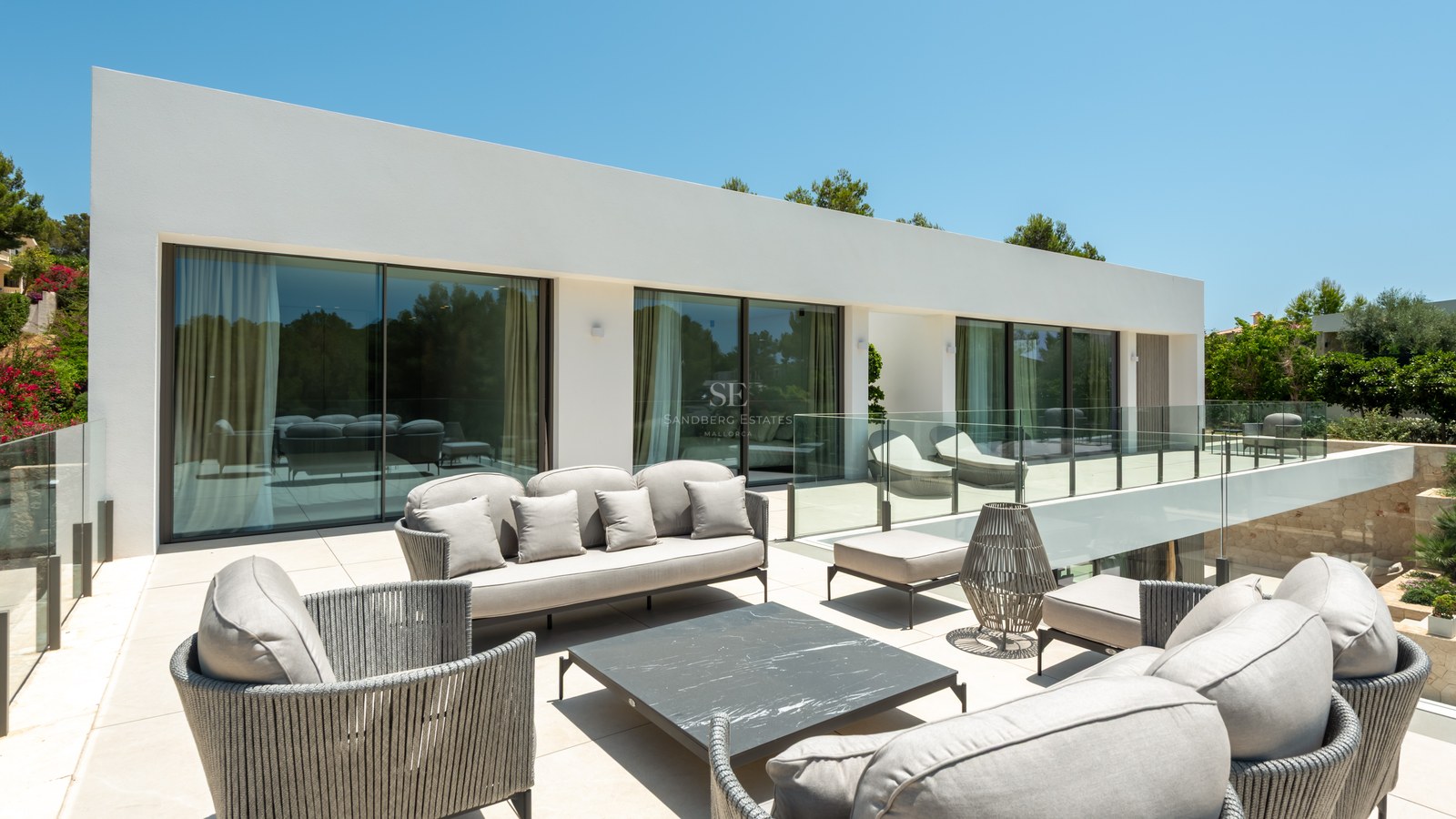 Terrasse de villa moderne avec salon de jardin gris, garde-corps en verre et larges baies vitrées.