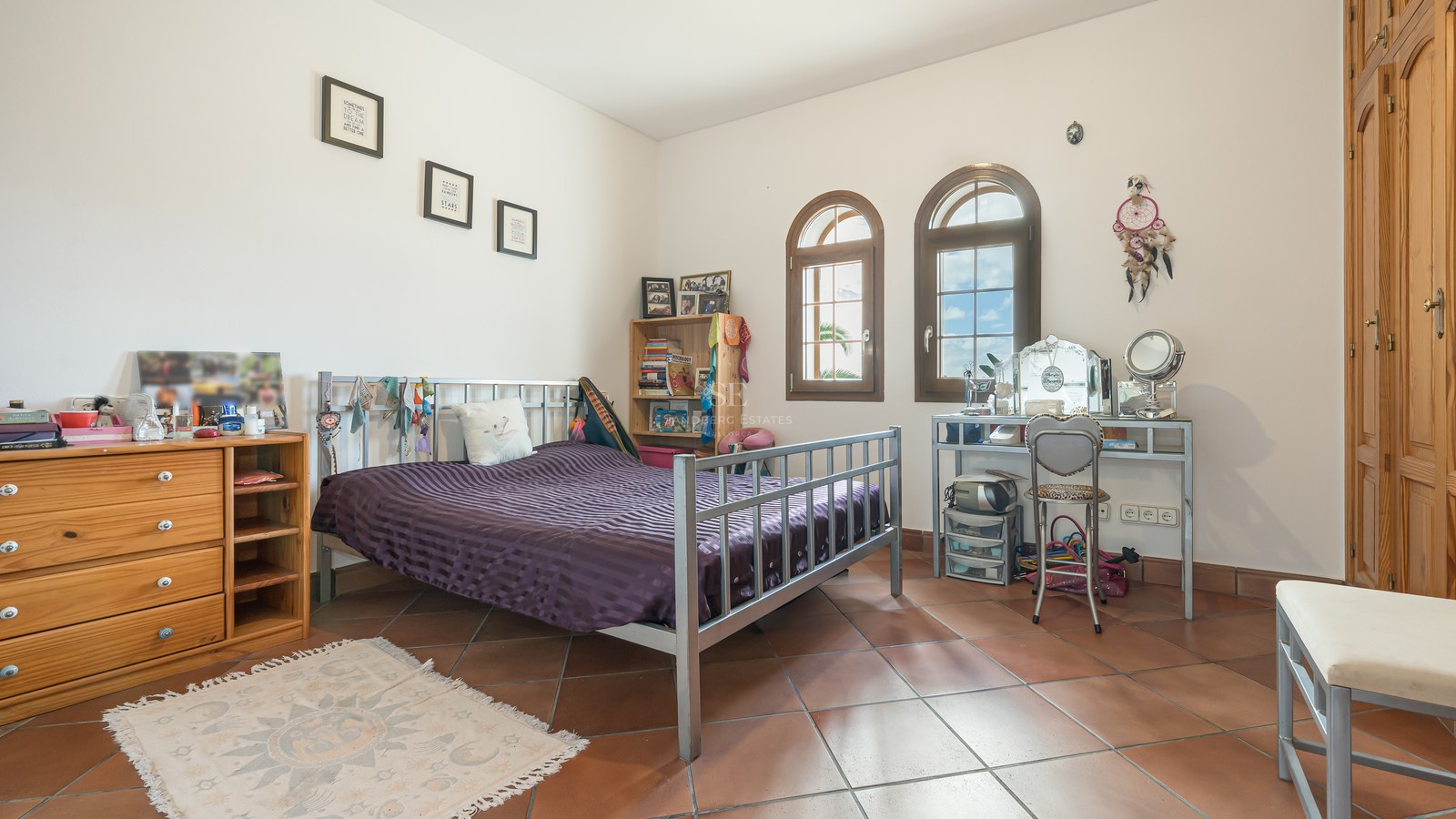 Chambre avec sol en terre cuite, lit en métal, fenêtres cintrées en bois et placards intégrés sous lumière naturelle.