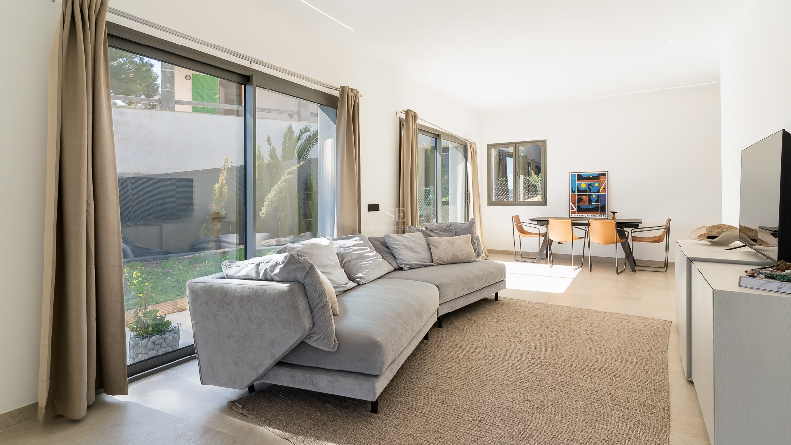 Helles Wohnzimmer mit grauem Sofa, bodentiefen Glasfronten zum Garten und Essbereich mit Lederstühlen.