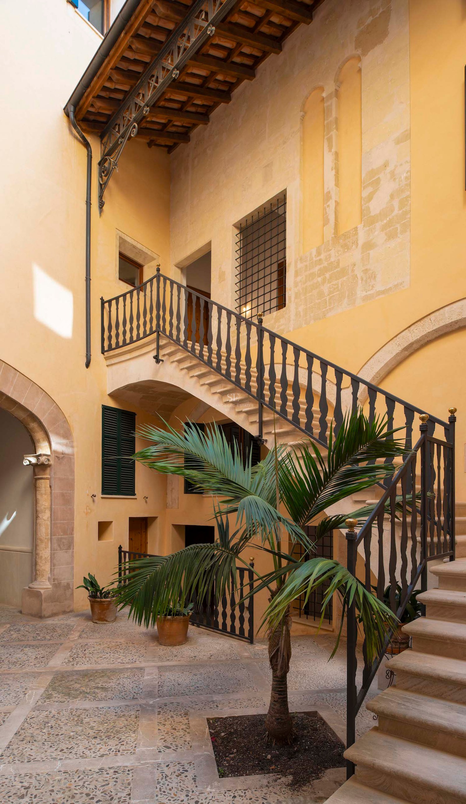 Traditional courtyard with yellow walls, a stone staircase with iron railings, and a central palm tree.