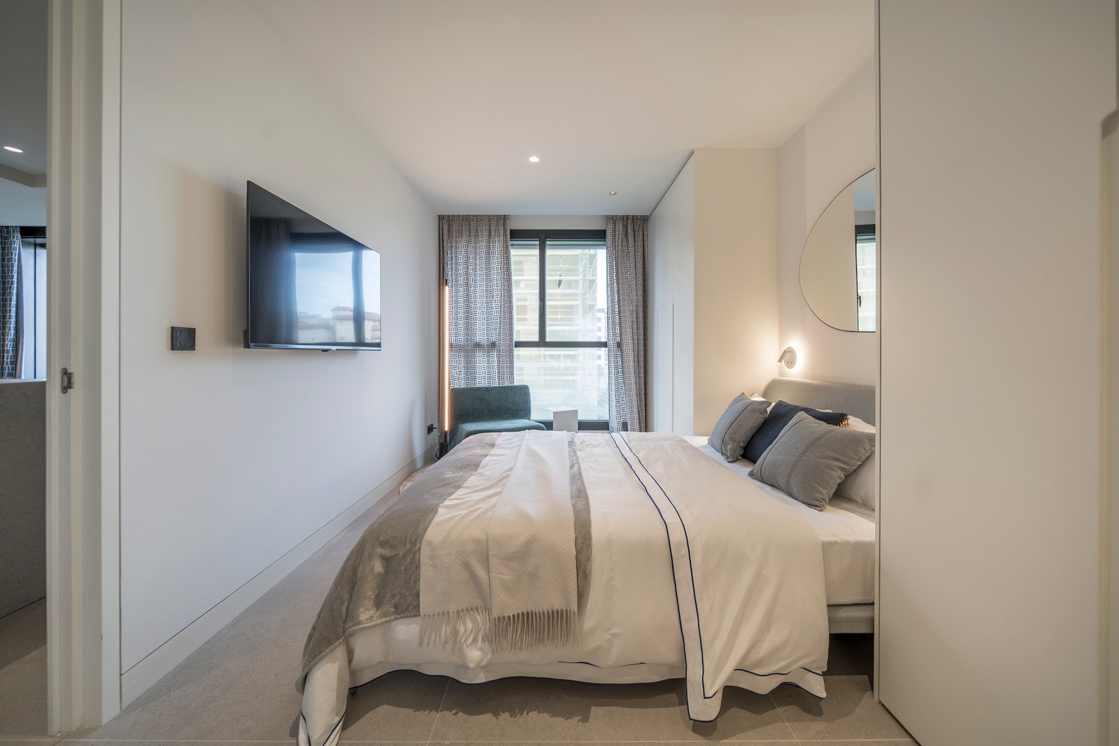 Chambre lumineuse avec TV murale, lit tapissé gris, miroir organique et grande fenêtre.