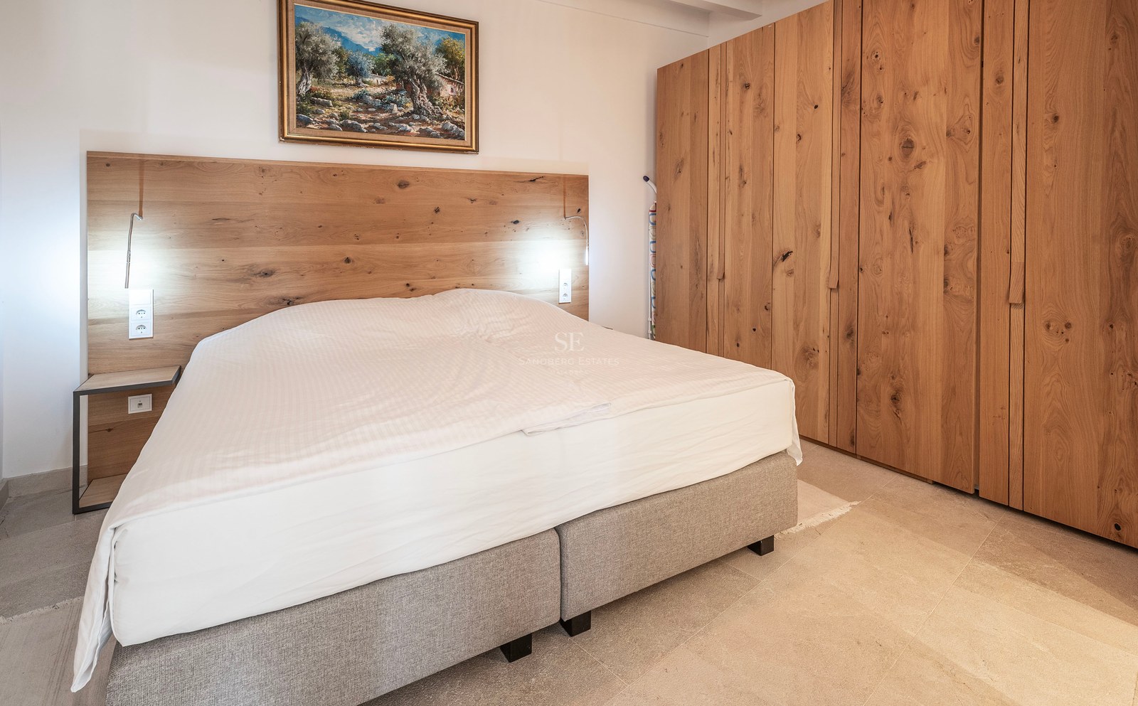 A modern bedroom featuring a large bed with a wooden headboard, extensive oak wardrobes, and a painting on the wall.