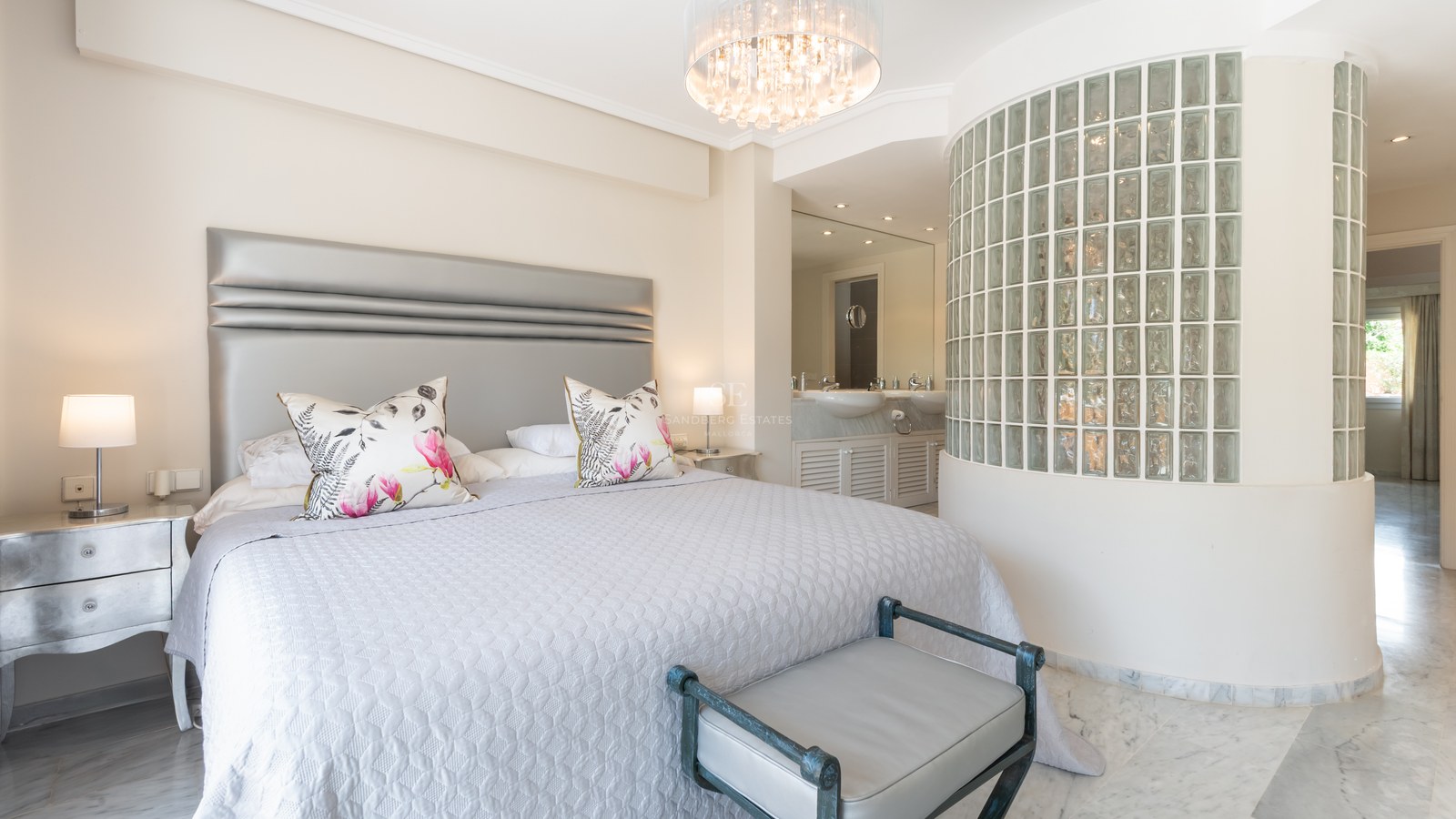 Chambre de luxe avec grand lit, sol en marbre, lustre en cristal et mur incurvé en briques de verre vers la salle de bain.