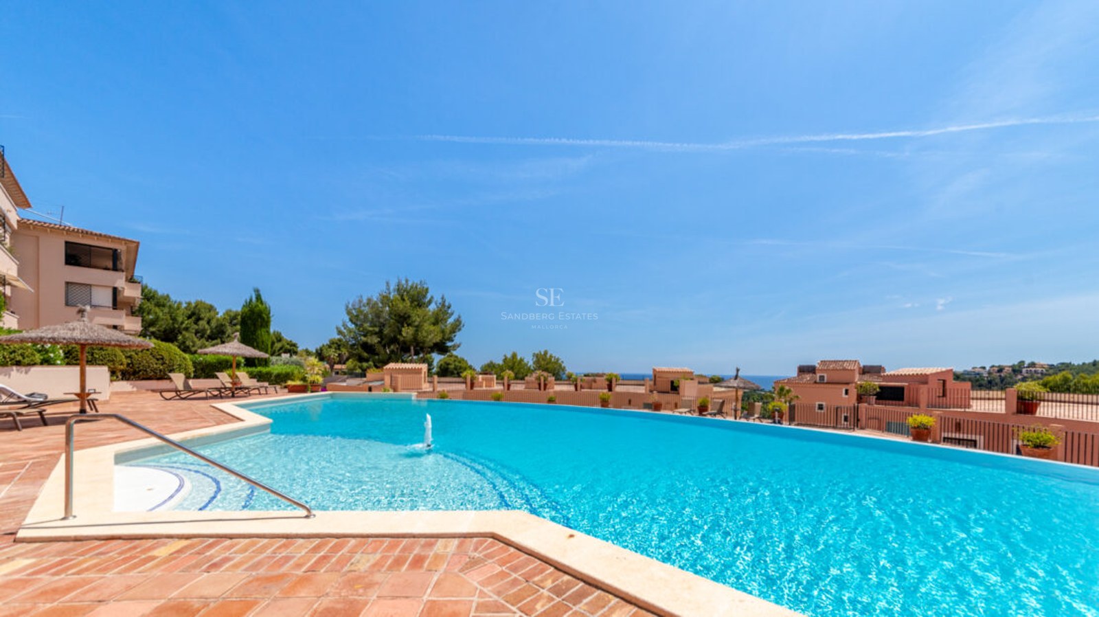 Grande piscine bleue incurvée avec fontaine, terrasse en terre cuite, parasols en paille et vue sur mer.