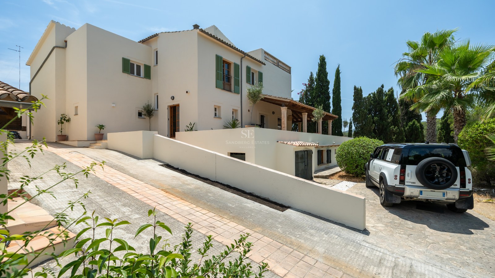 Exterior view of a white multi-level Mediterranean villa with green shutters, a paved driveway, and palm trees.