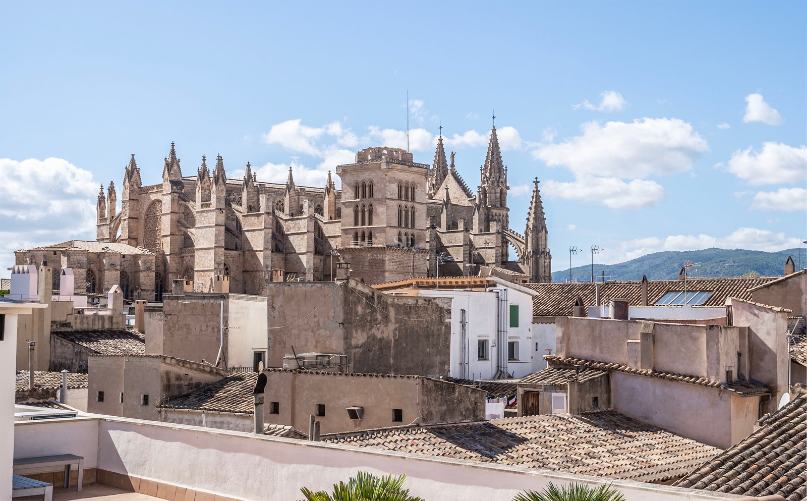 A large Gothic cathedral towers over the terracotta rooftops of a Mediterranean city under a blue sky.