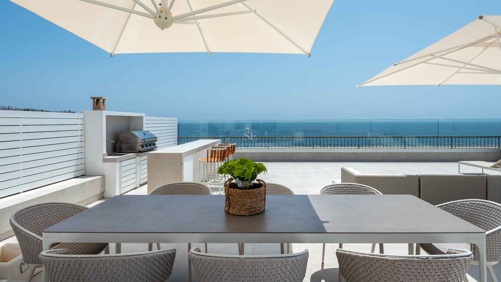 Luxury rooftop terrace featuring a dining area, outdoor kitchen, and glass railings overlooking the blue Mediterranean sea.