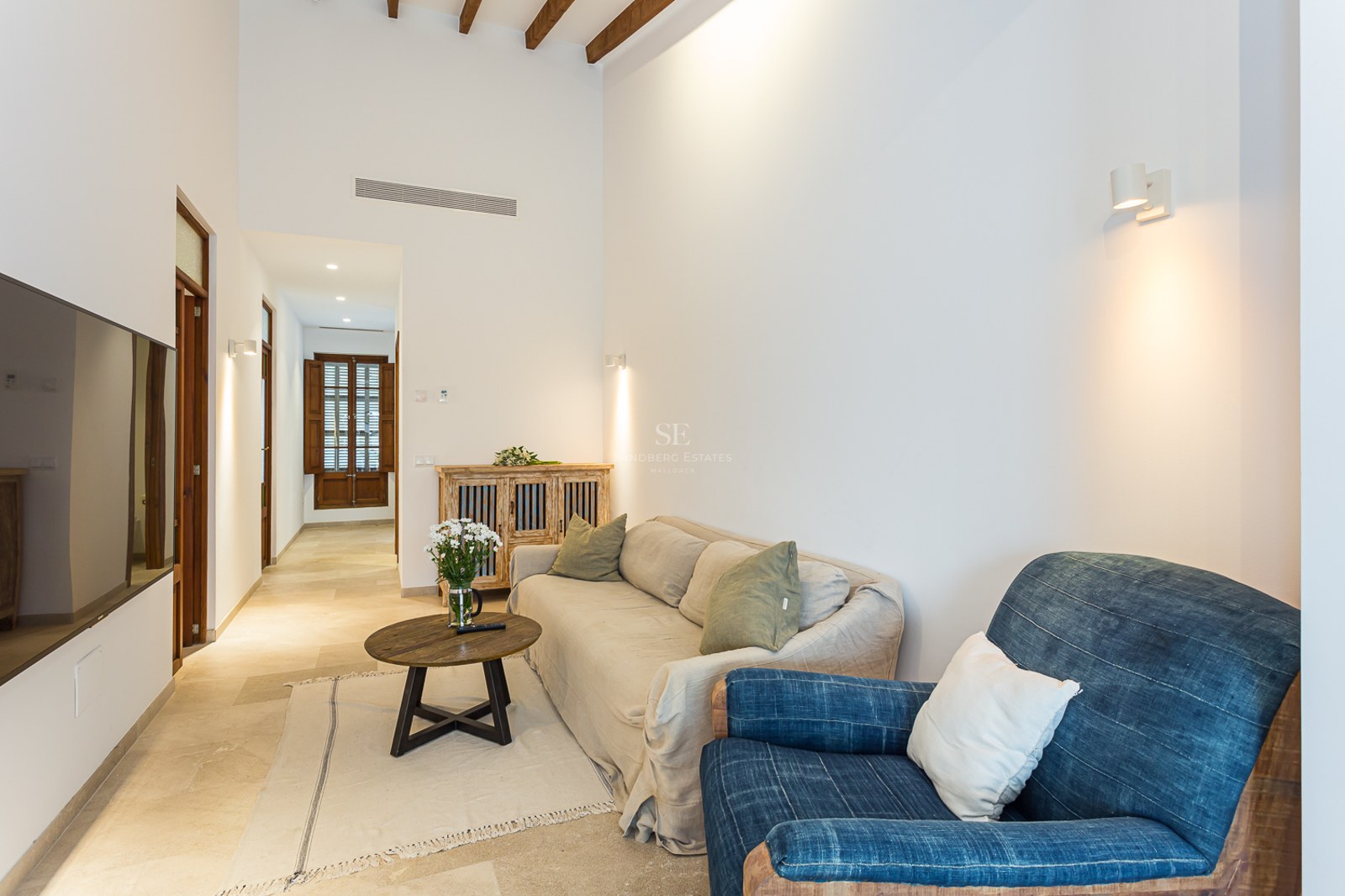 Salón luminoso con paredes blancas, vigas de madera en el techo, sofá beige, sillón azul y mesa de centro redonda.