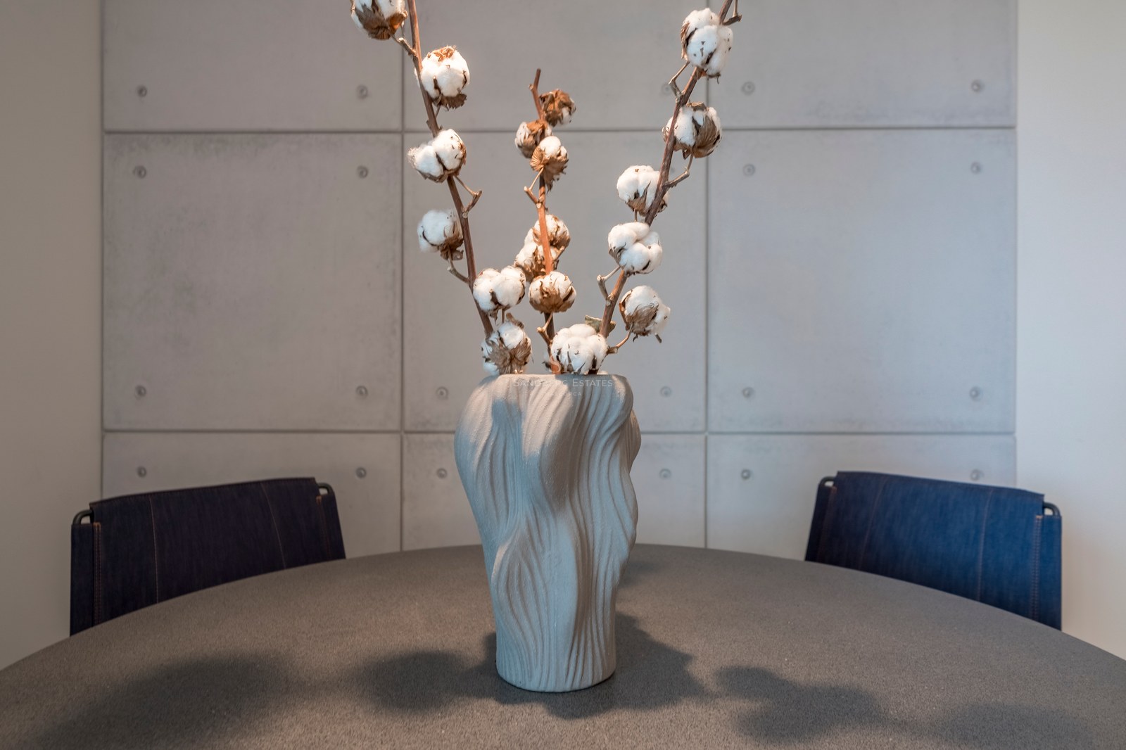 Vase gris texturé avec des branches de coton sur une table sombre devant un mur en panneaux de béton.