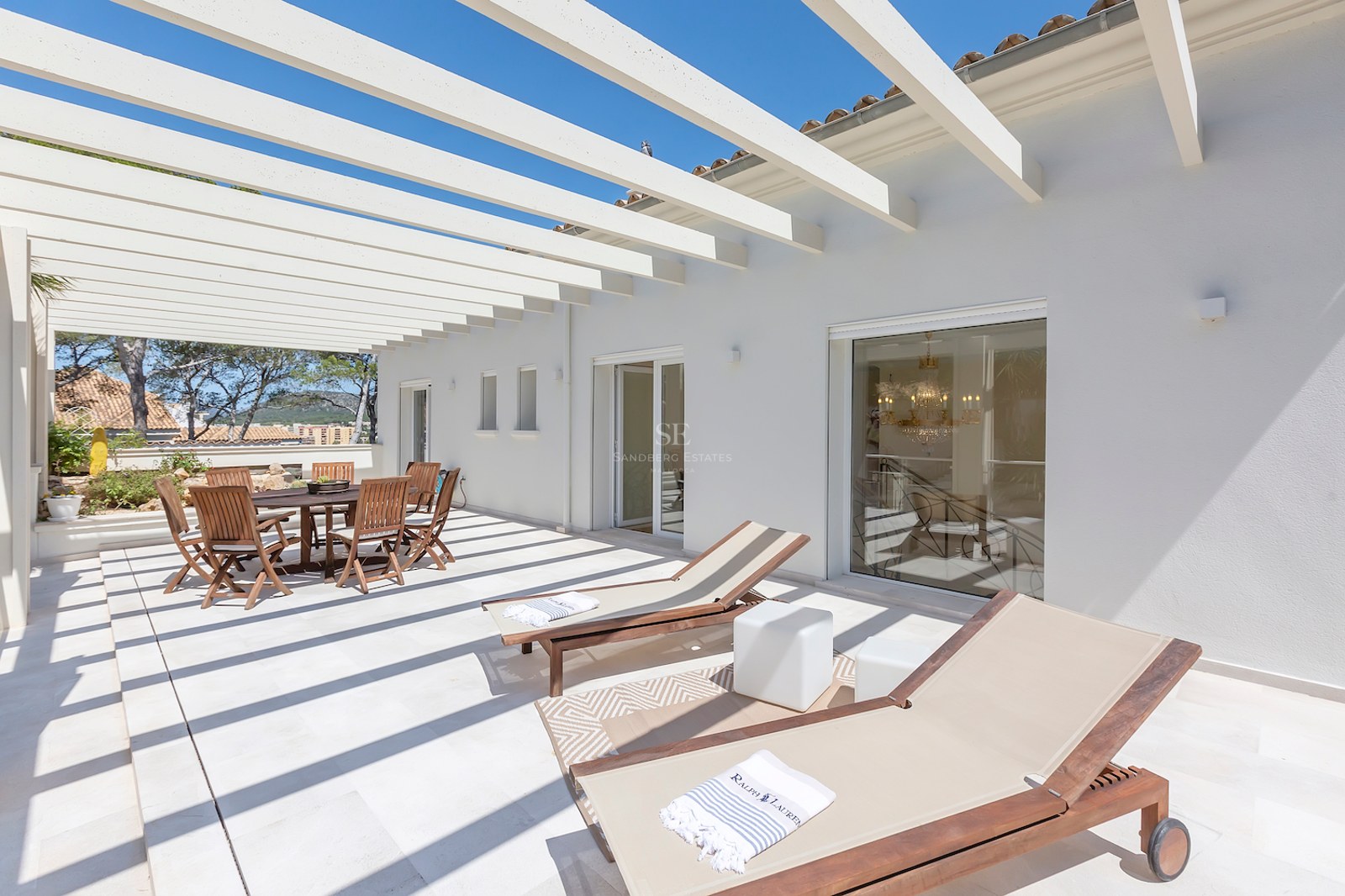 Bright white terrace with a wooden pergola, outdoor dining table, and sun loungers under a clear blue sky.