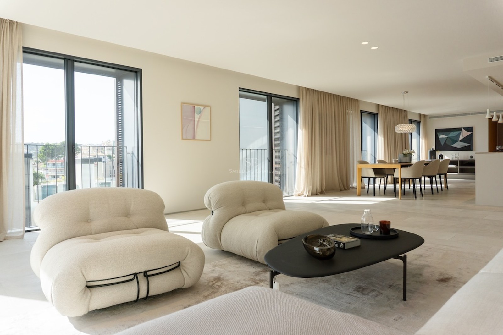 Ljust vardagsrum med designfåtöljer i beige, svart soffbord och matplats intill stora fönsterpartier.