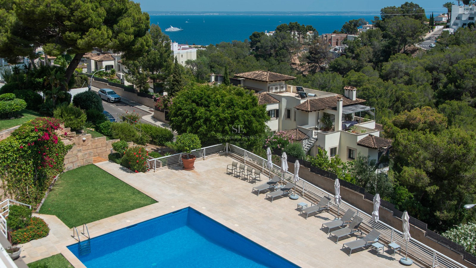 Flygvy över en blå pool och solterrass med solstolar med utsikt över Medelhavet och villor.
