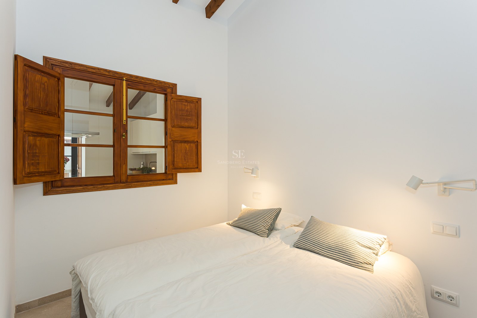 Dormitorio luminoso con cama blanca, cojines a rayas, ventana de madera con contraventanas y vigas de madera vistas.