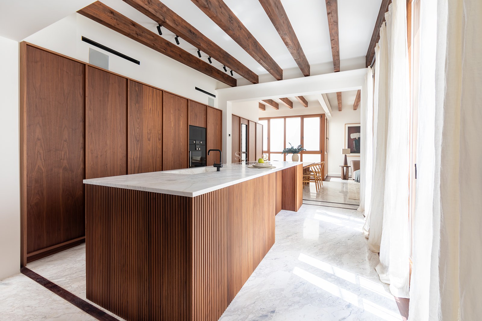 Modern kitchen with a fluted wood island, marble countertops, exposed ceiling beams, and natural light from large windows.
