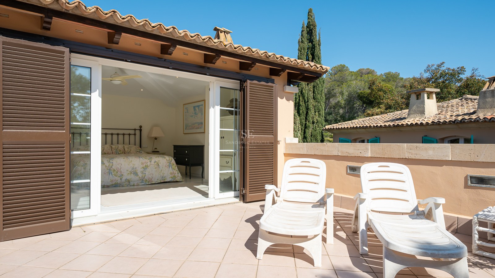 Chambre avec lit en fer forgé s'ouvrant sur une grande terrasse en terre cuite avec deux chaises longues.