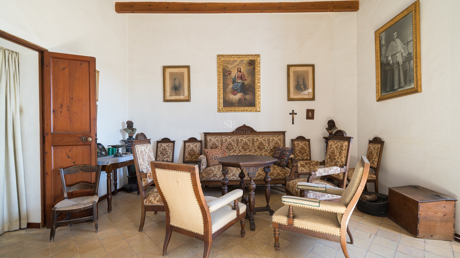 Sala de estar clásica con muebles de madera antiguos, tapicería estampada, pinturas religiosas y suelo de baldosas.