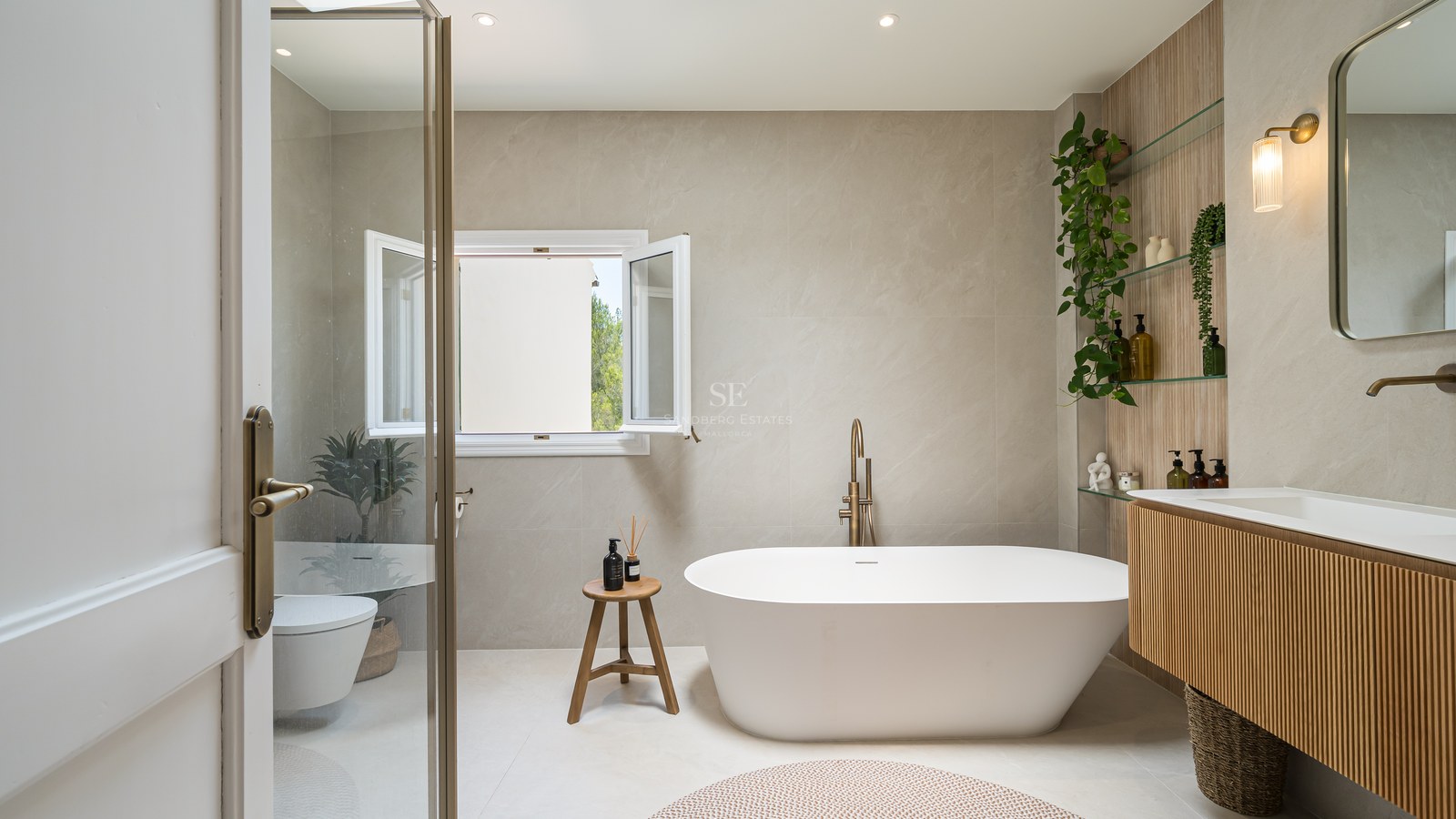 Modern bathroom featuring a white freestanding bathtub, brushed brass fixtures, stone-tiled walls, and an oak vanity.