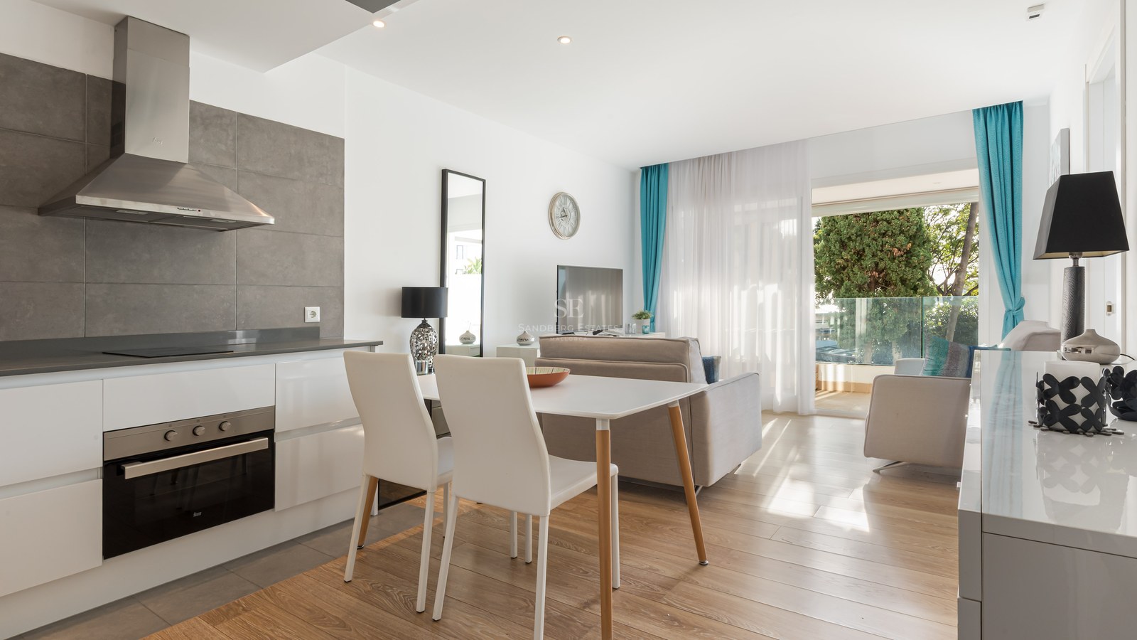 Bright open-plan room featuring a kitchen, dining area, and lounge leading to a sunny balcony with trees.