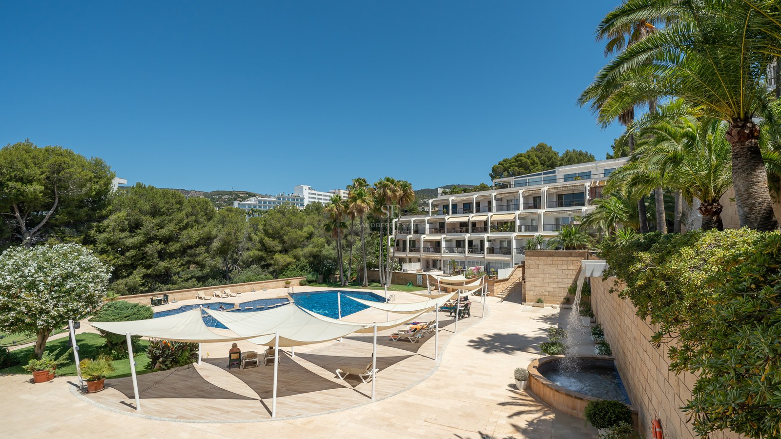 A large blue swimming pool surrounded by a stone terrace with white shade sails and palm trees under a clear blue sky.