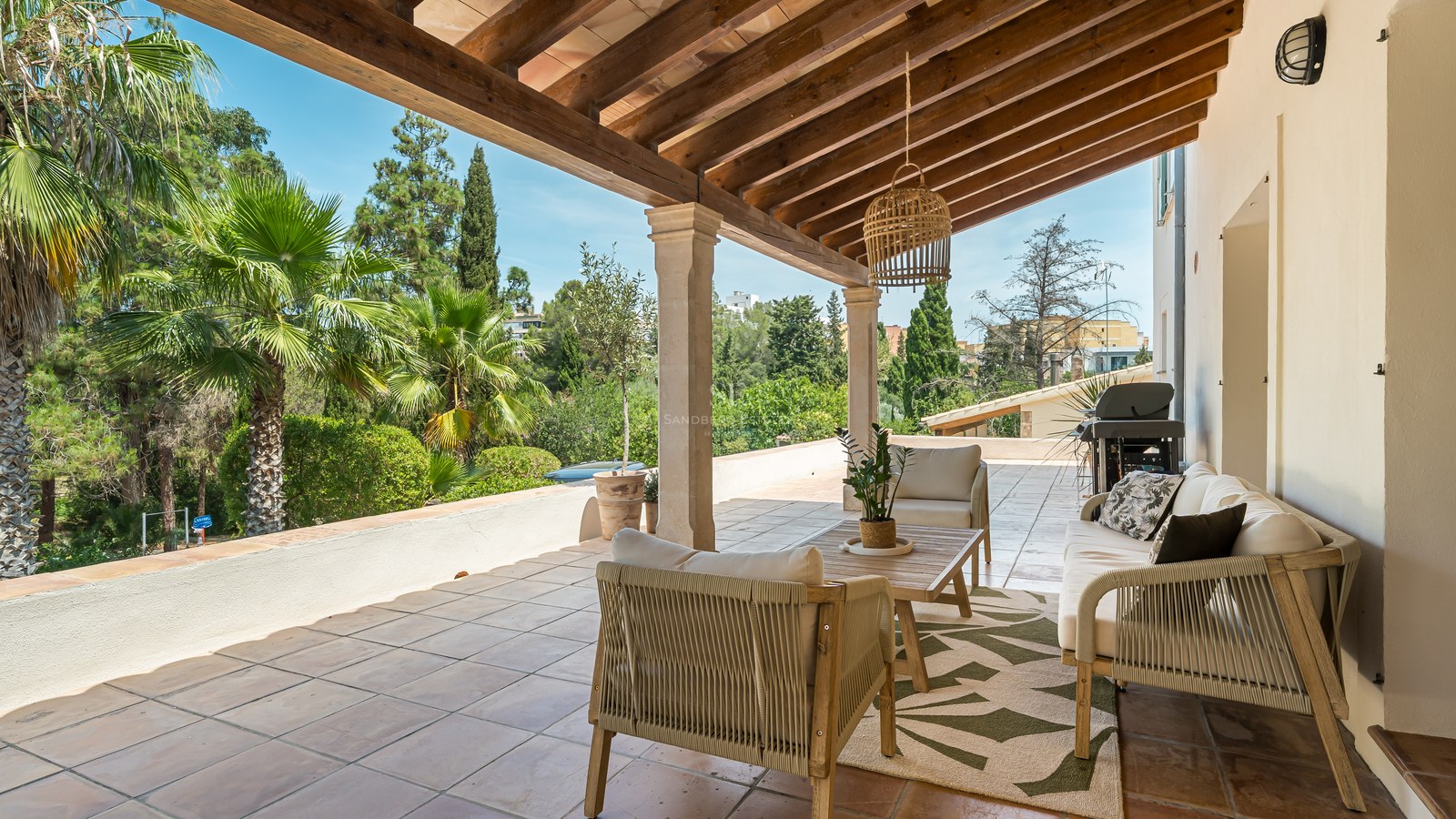 Mediterranean terrace with wooden beam ceiling, terracotta floor, outdoor lounge furniture, and views of palm trees.