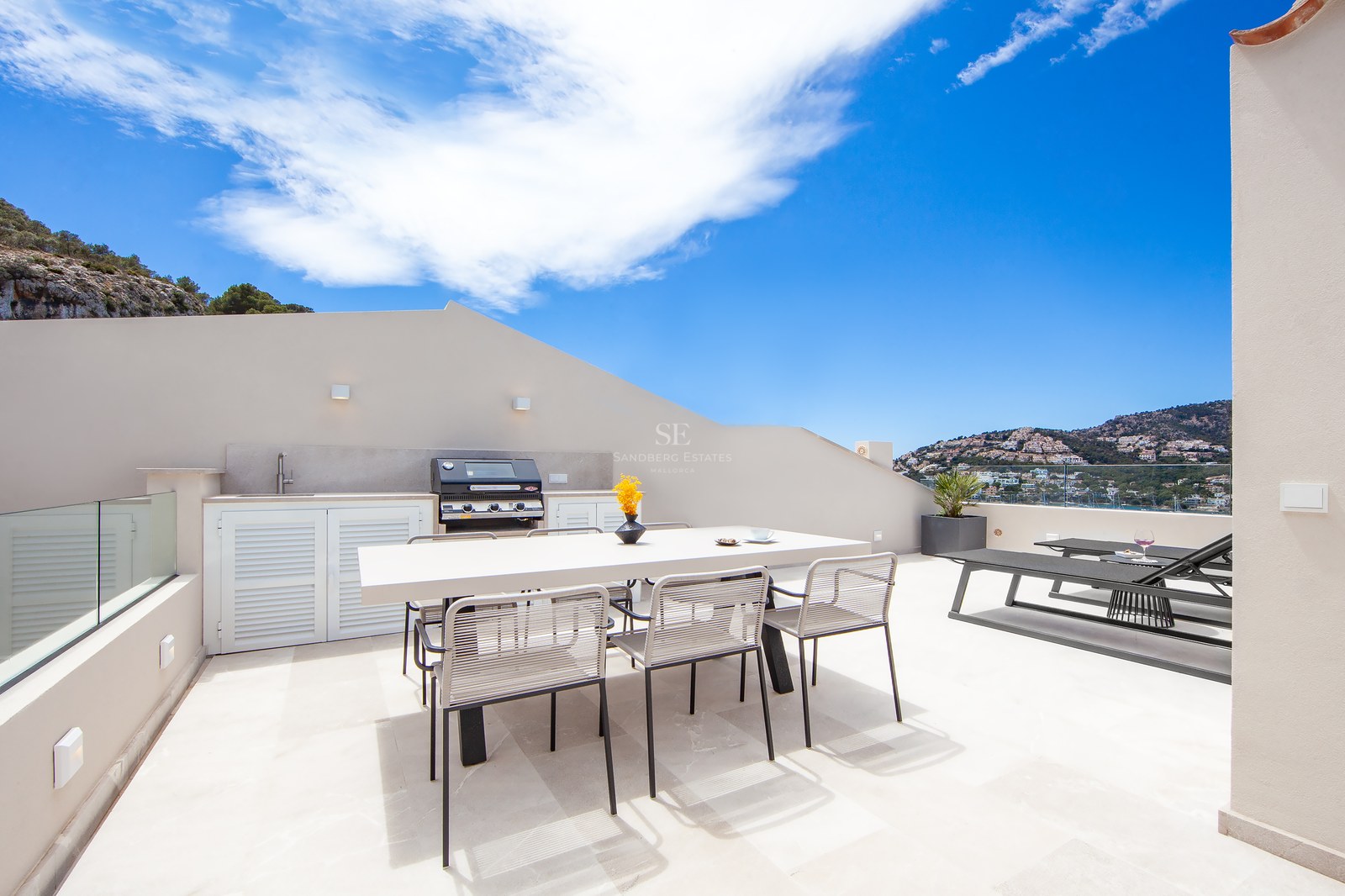 Modern rooftop terrace featuring an outdoor kitchen, dining table, and sun loungers with panoramic coastal views.