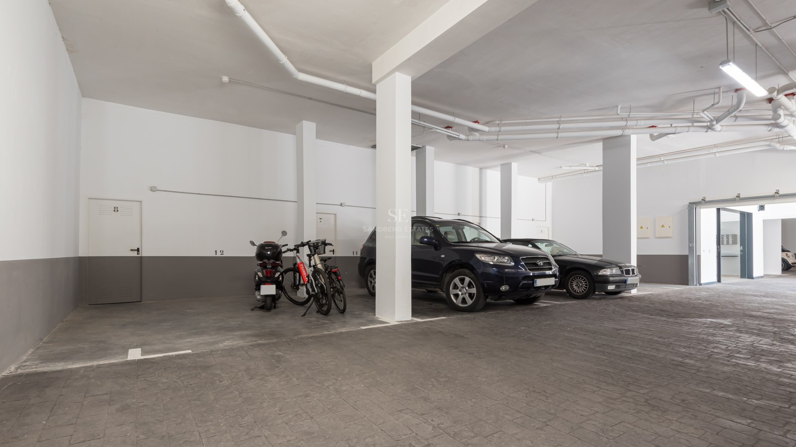 Underground parking garage with white pillars, grey textured stone flooring, and cars parked under artificial lighting.