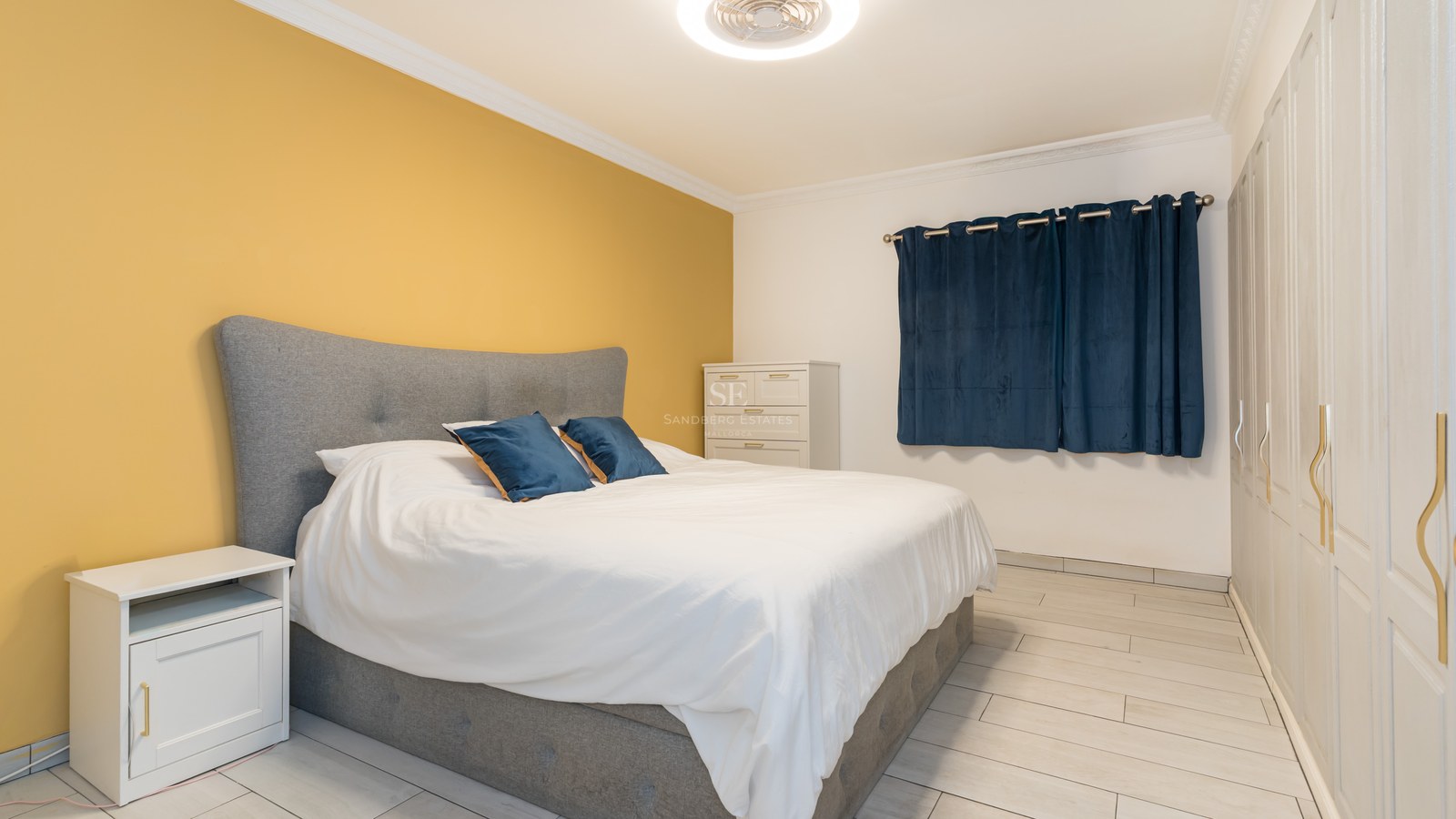 A bright bedroom featuring a gray bed, yellow accent wall, white built-in wardrobes, and dark blue curtains.