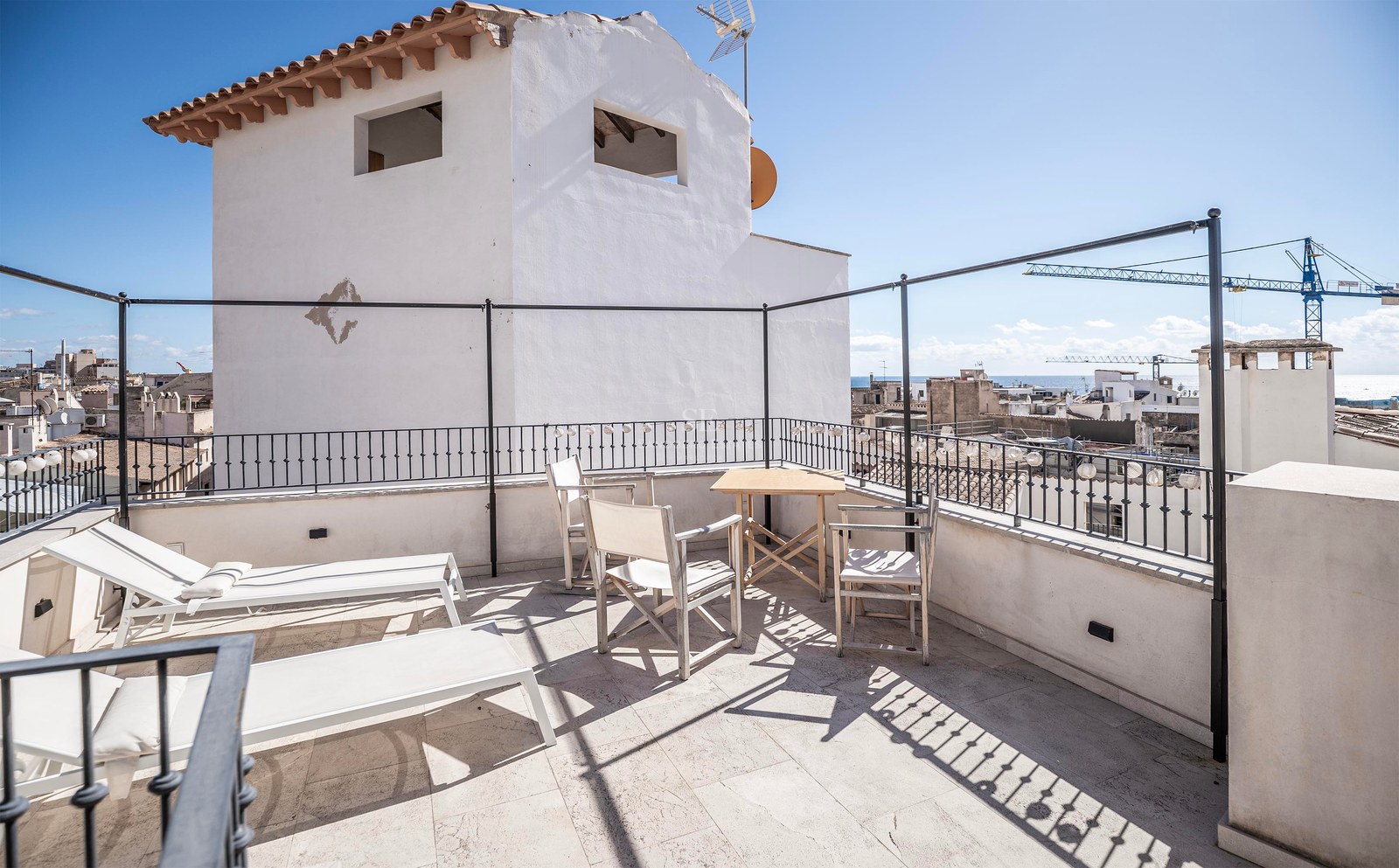 Sunny rooftop terrace featuring white lounge chairs, a wooden table, and distant sea views over city rooftops.