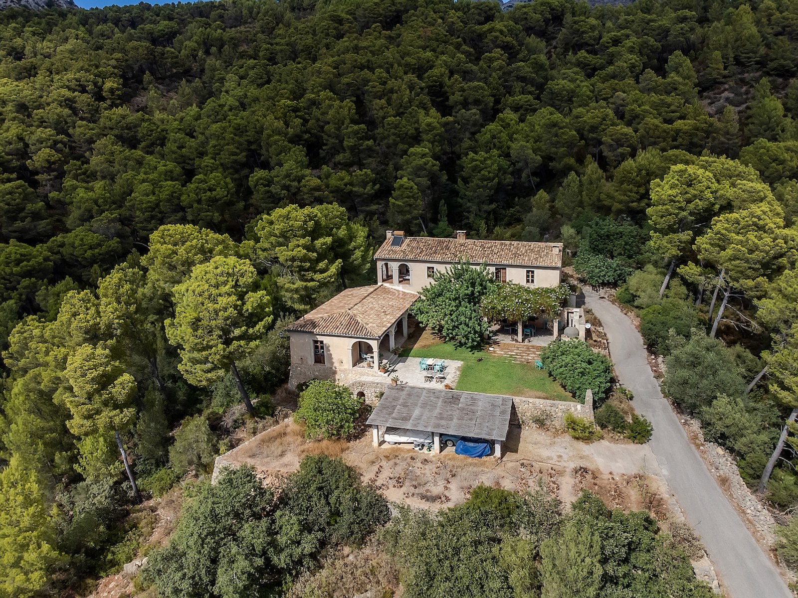 Vue d'oiseau d'une villa rustique avec toits en terre cuite entourée d'une forêt de pins dense sur une colline.