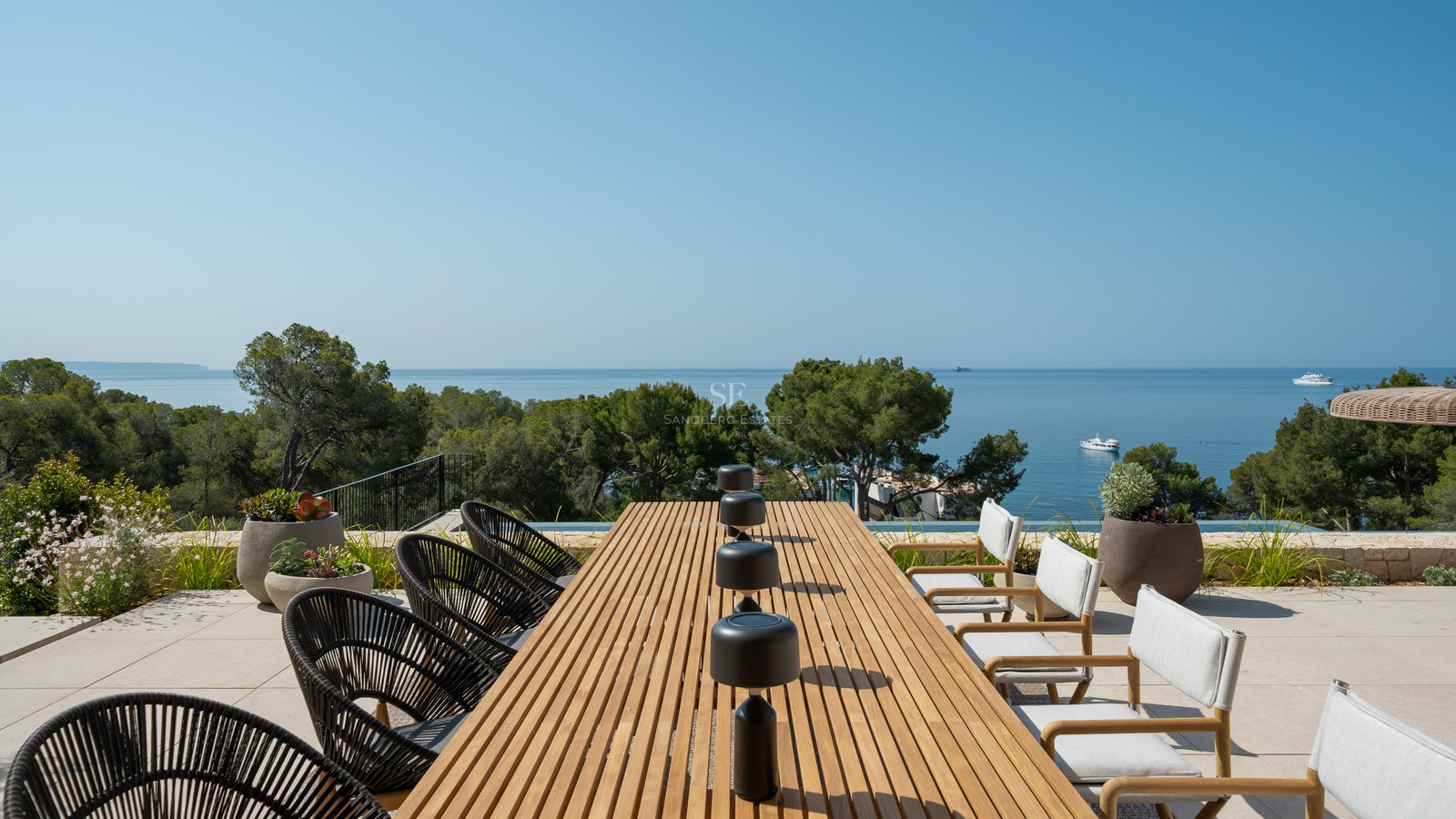 A long wooden dining table on a sunny stone terrace overlooking the Mediterranean Sea and green pine trees.