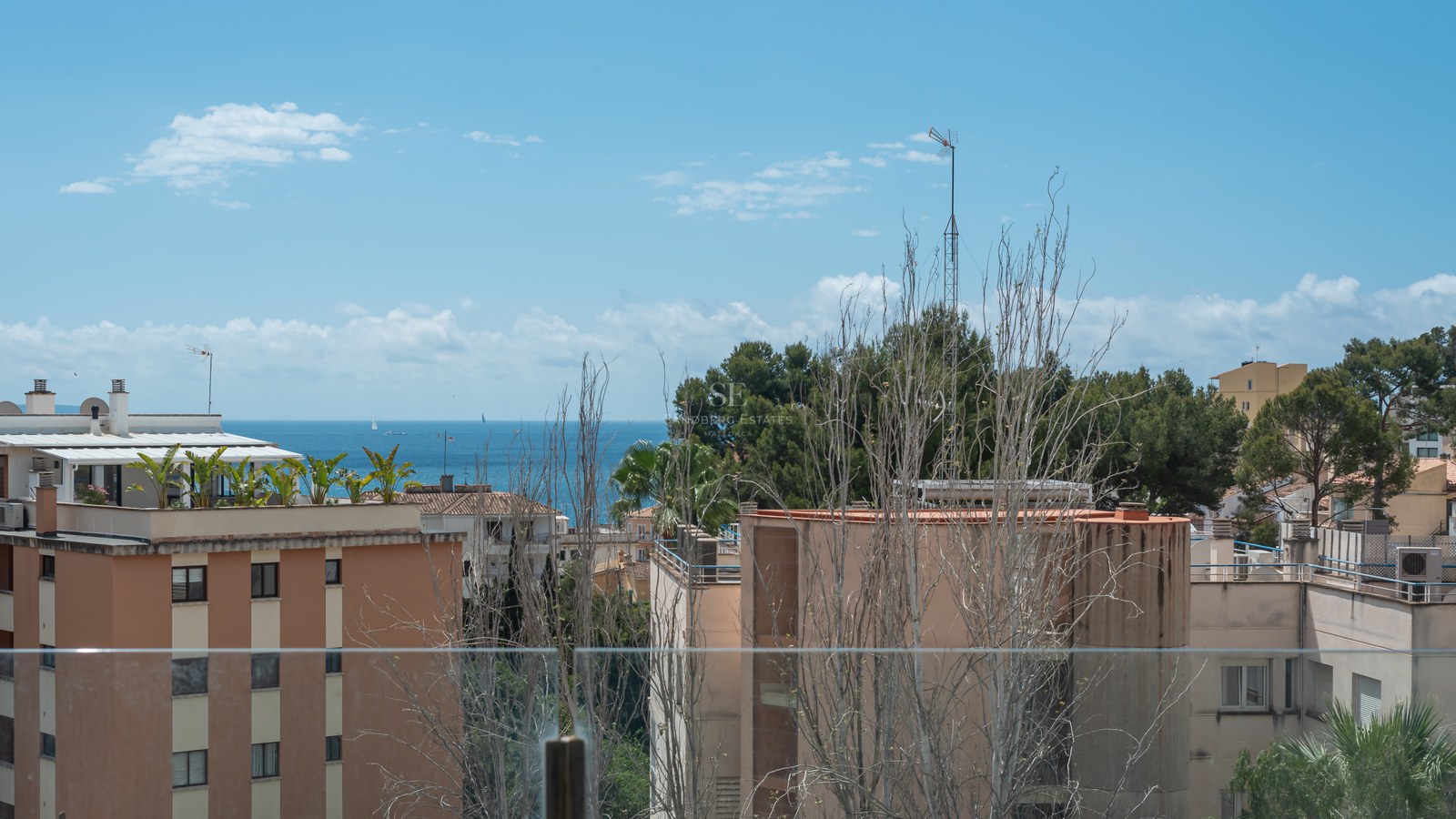 View of the blue Mediterranean sea and coastal buildings through a clear glass balcony railing under a blue sky.