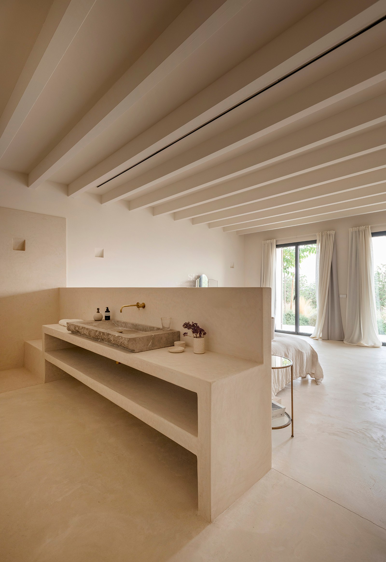 Minimalist bedroom with a built-in stone washbasin, exposed ceiling beams, and floor-to-ceiling glass doors.