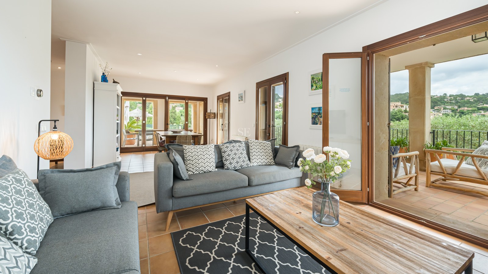 Modern grå soffa och soffbord i trä i ett ljust vardagsrum med terrakottagolv och stora glasdörrar.