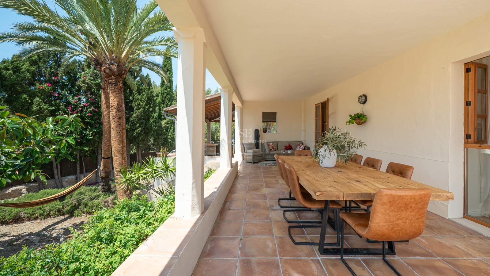 A covered terrace with a long wooden dining table, cognac leather chairs, terracotta floors, and lush garden views.