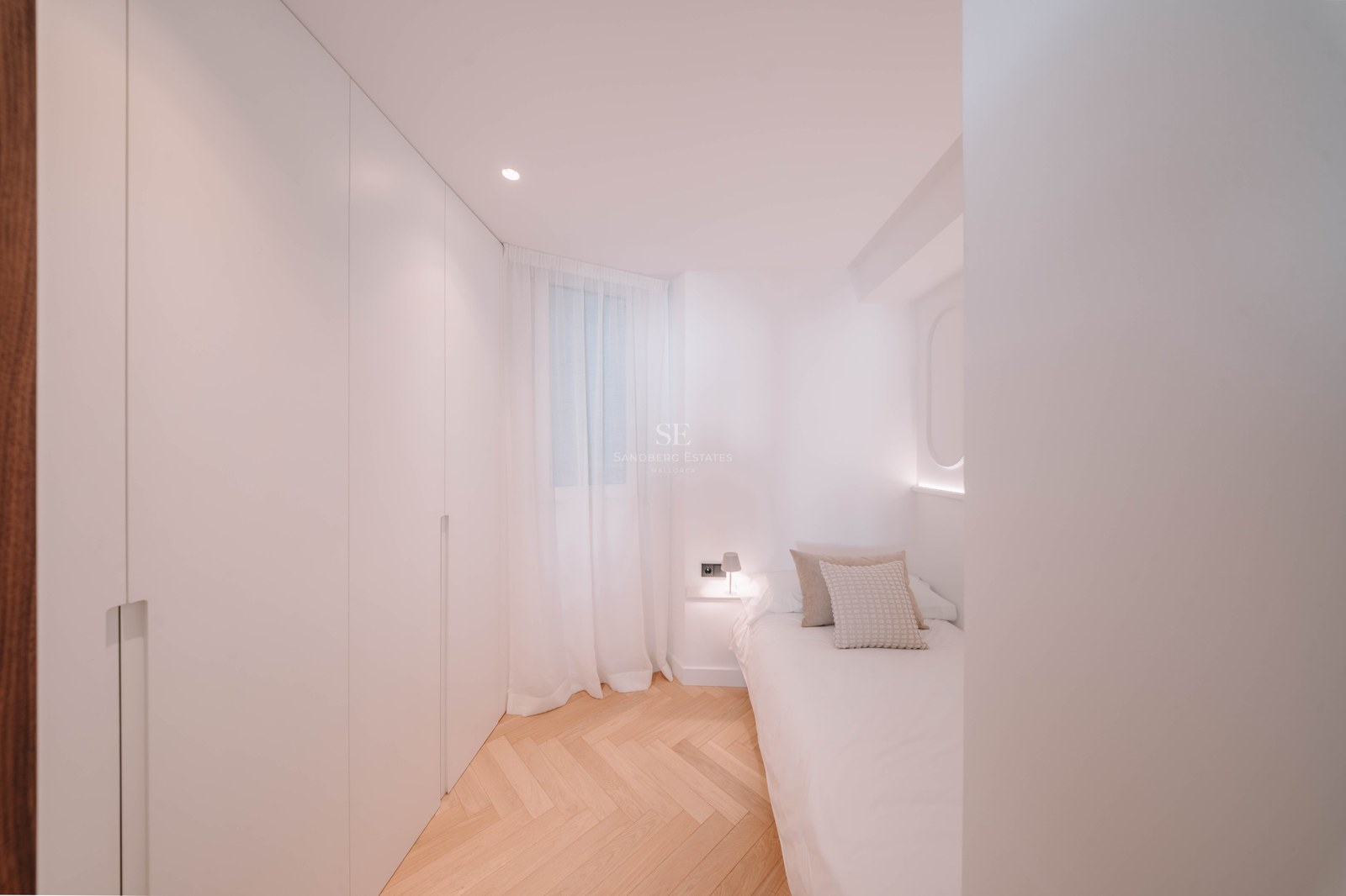 Chambre lumineuse avec placards blancs intégrés, parquet en chevrons et un lit simple avec éclairage doux.