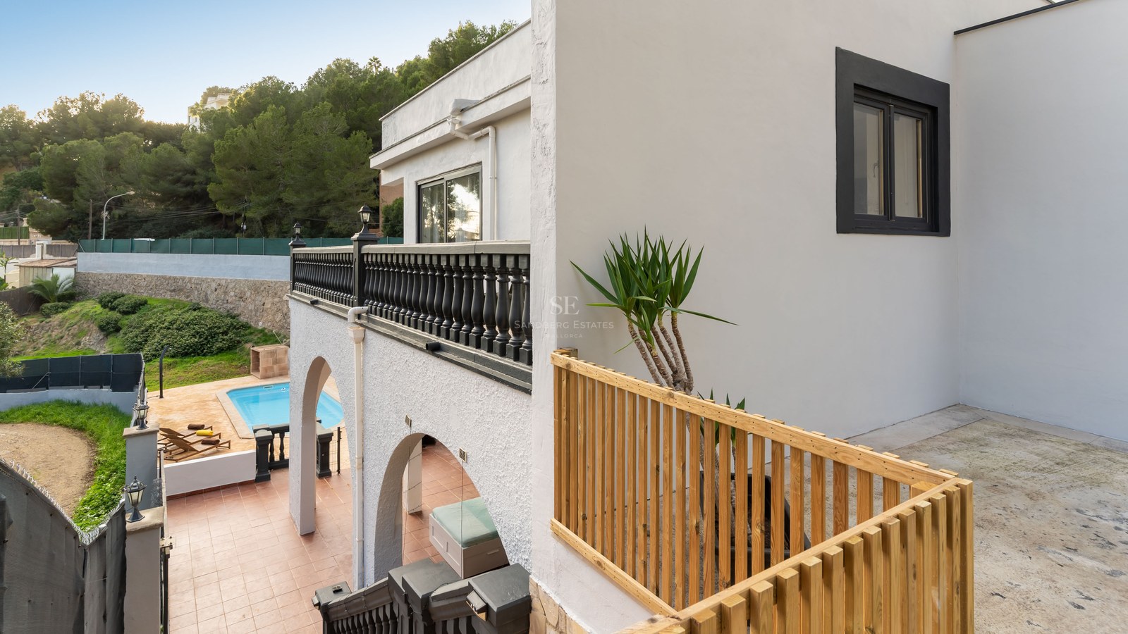 White multi-level villa with black and wooden railings, overlooking a turquoise pool and hillside forest.