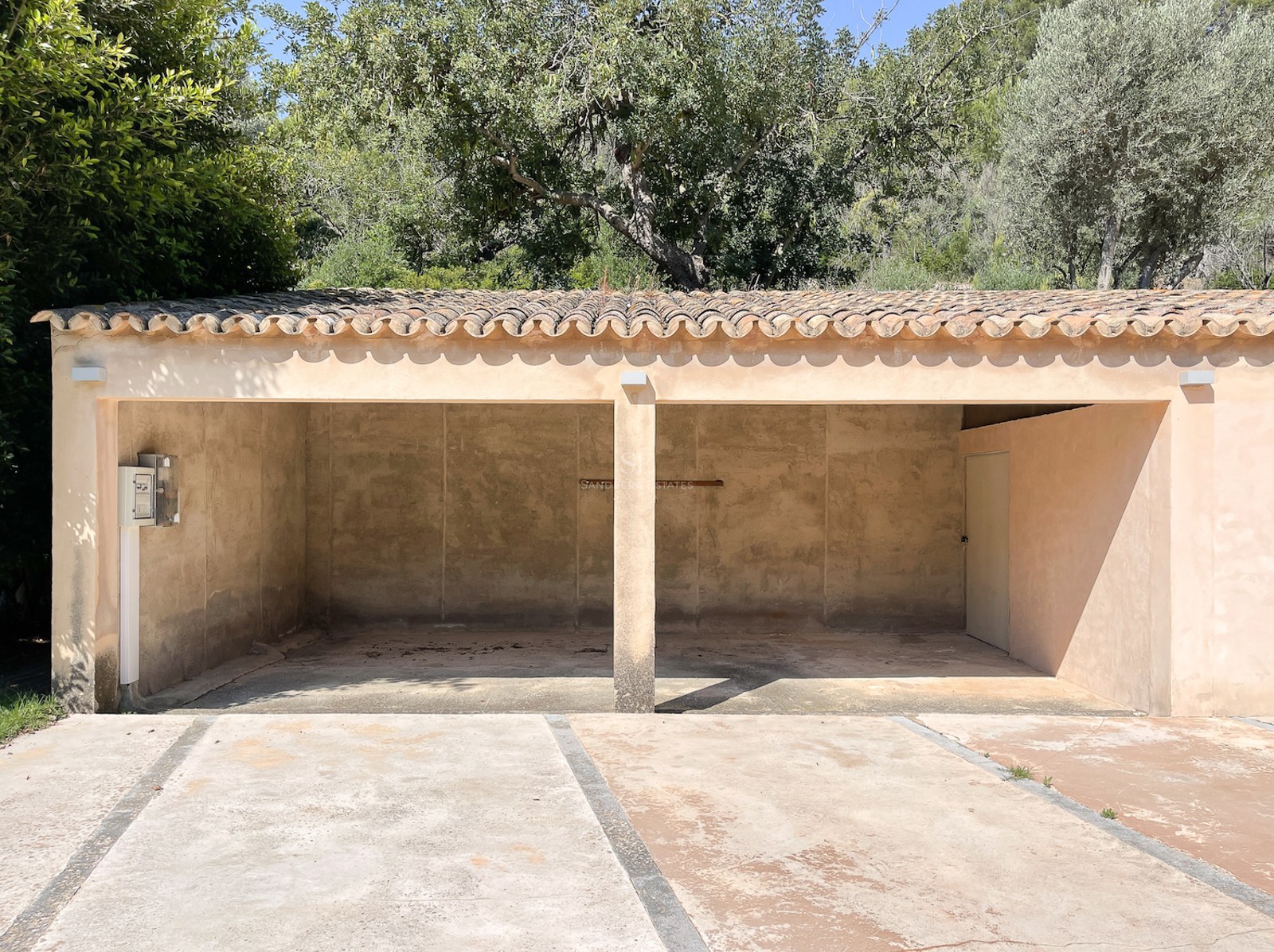 Garaje abierto de dos plazas con techo de tejas de terracota y paredes de color beige rodeado de vegetación.