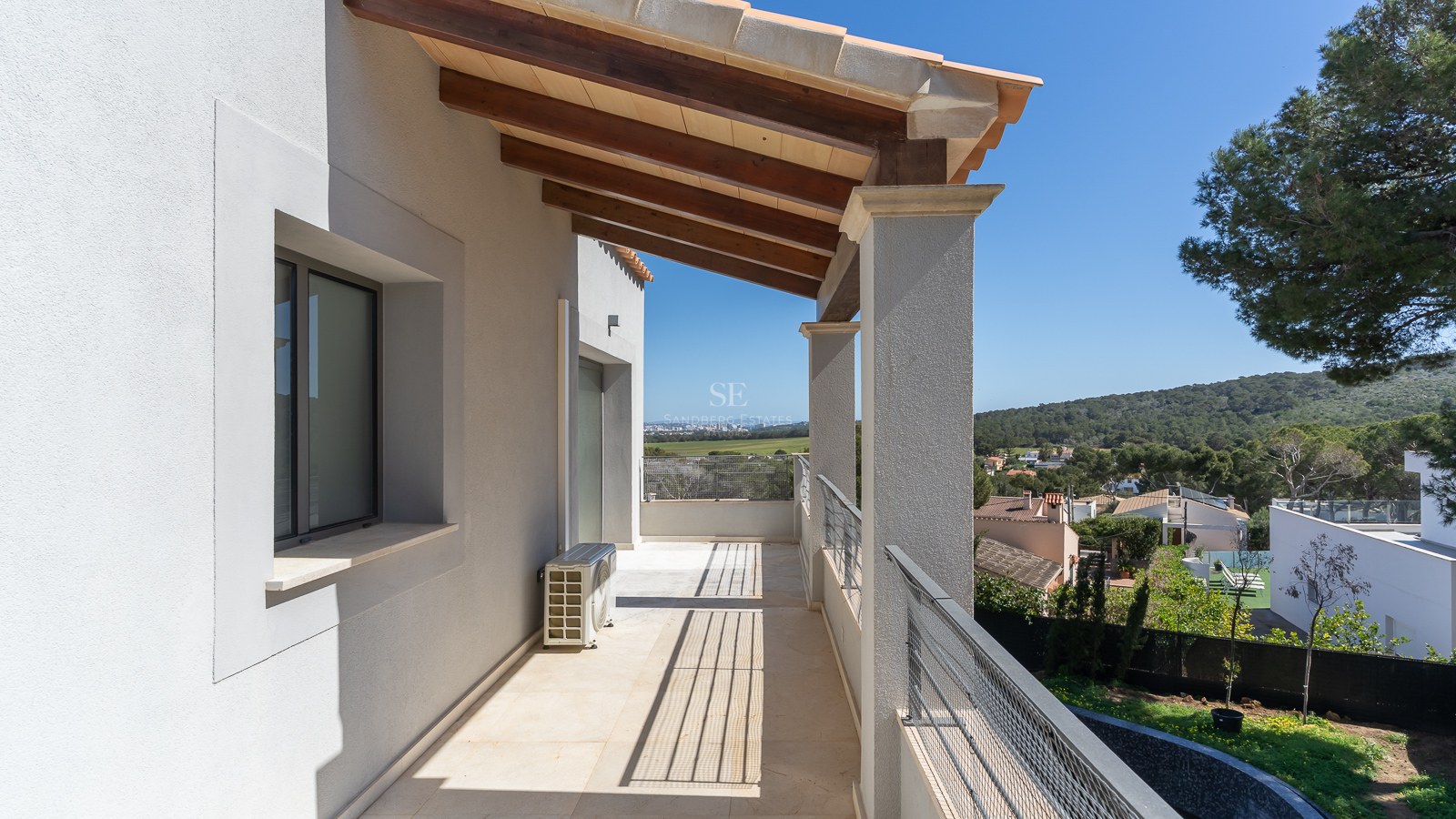Langer überdachter Balkon mit Holzbalken, weißen Wänden und Metallgeländer mit Blick auf ein grünes Tal.