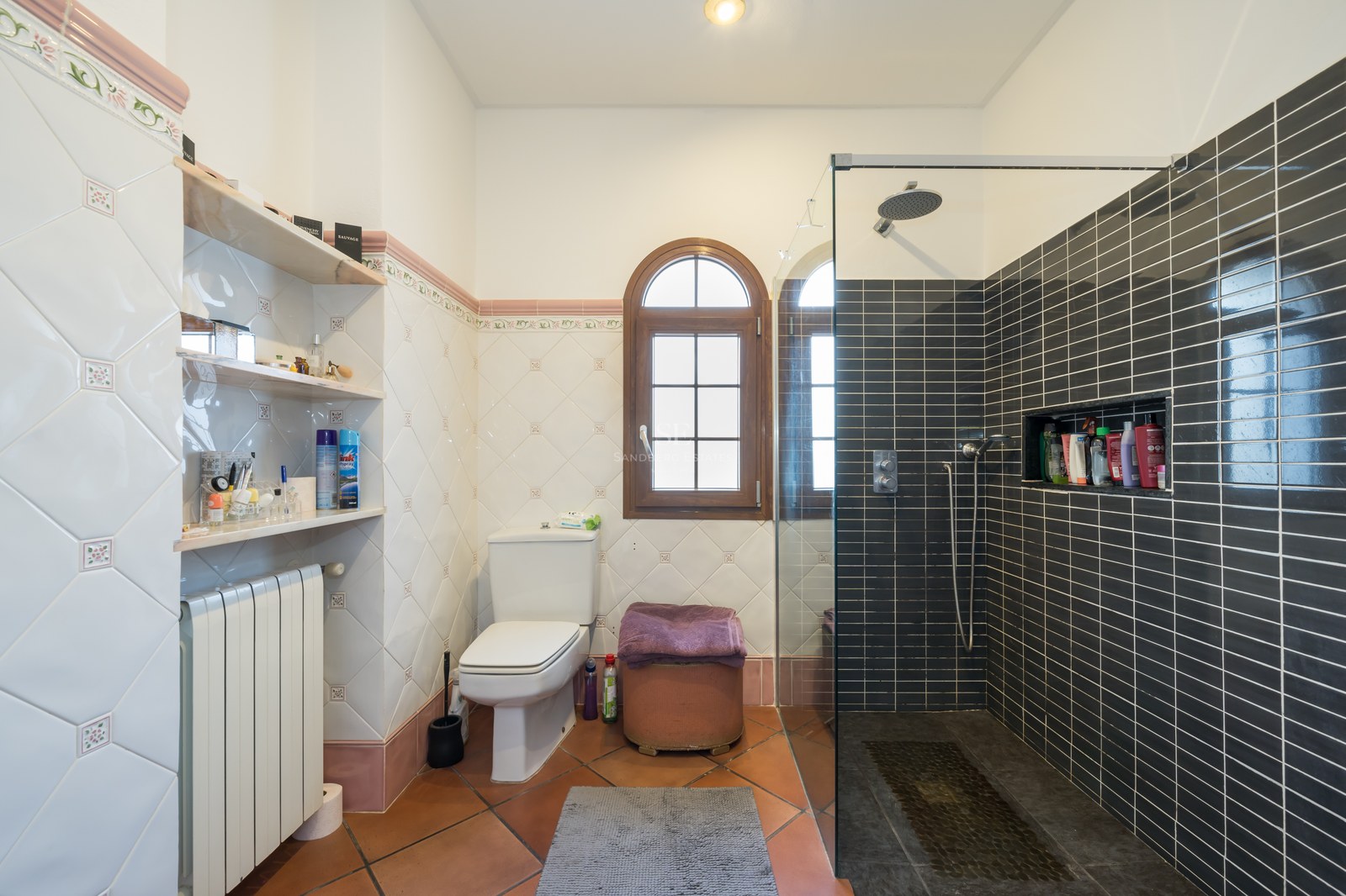 Salle de bain avec sol en terre cuite, carrelage décoratif, fenêtre cintrée en bois et douche moderne vitrée.
