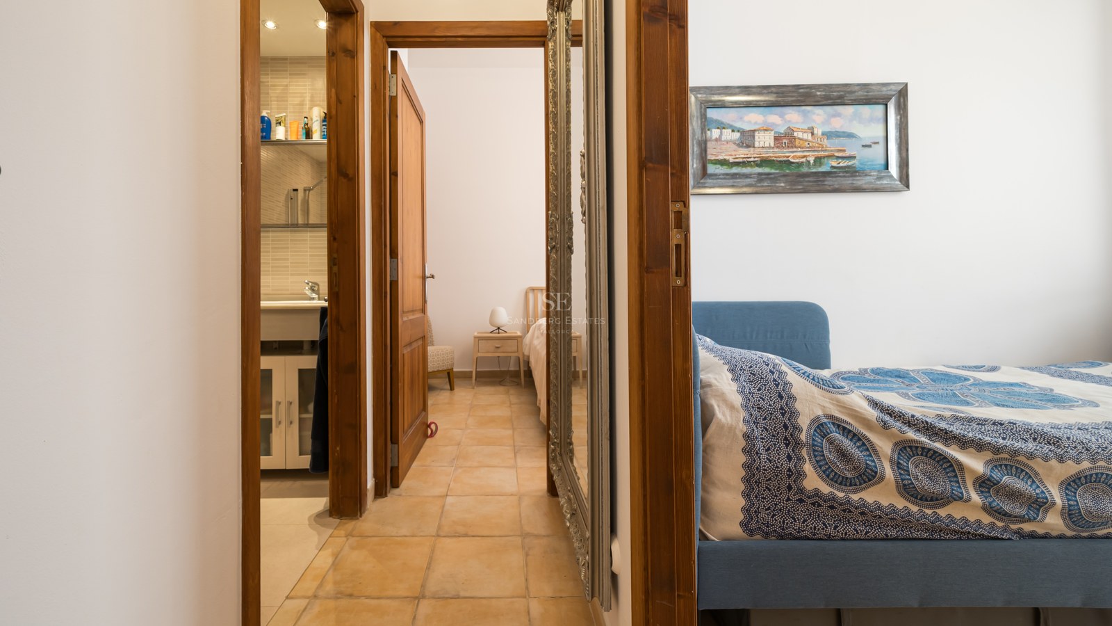 Un couloir avec carrelage en terre cuite menant à une salle de bain et deux chambres, doté d'un grand miroir argenté.